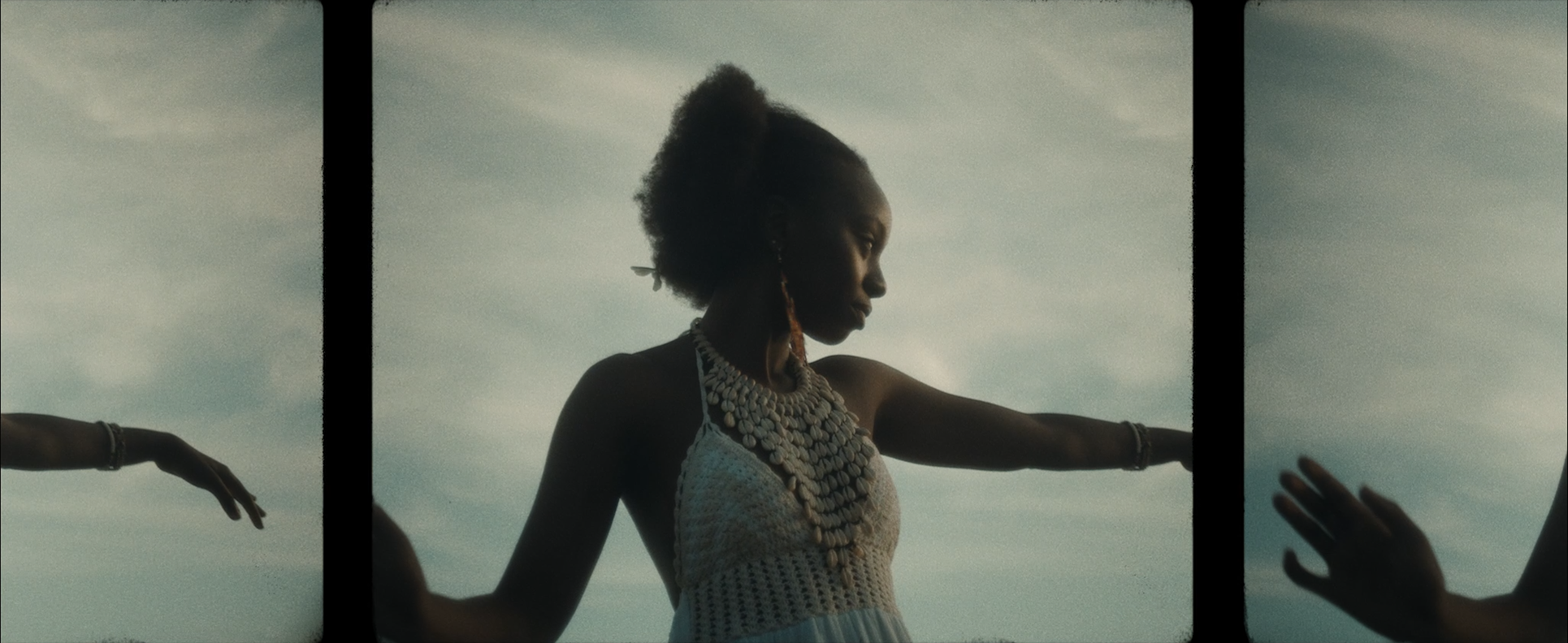 A woman dressed in a white crocheted top with layered necklaces, standing outdoors with arms extended, against a cloudy sky.