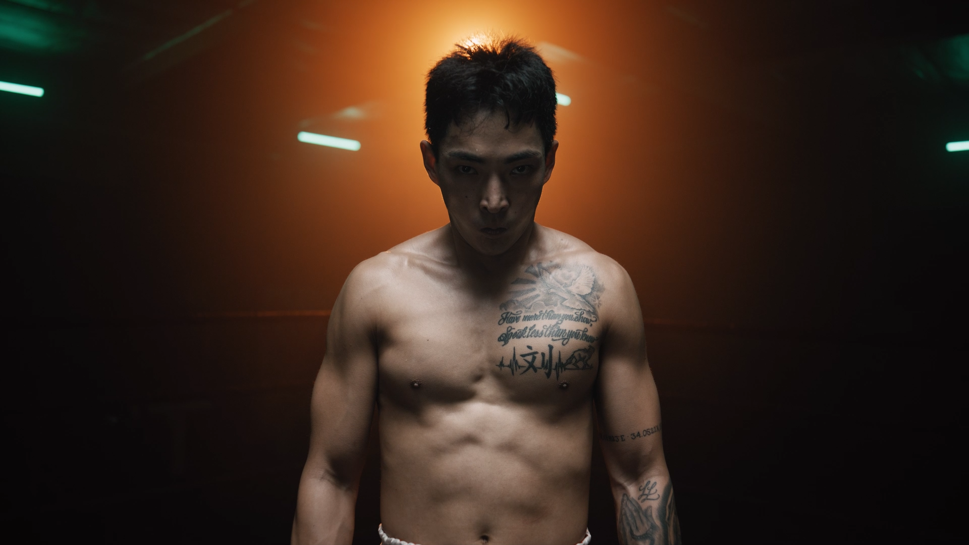 A shirtless man with tattooed chest and arms, standing in a dimly lit environment with orange and green lights.
