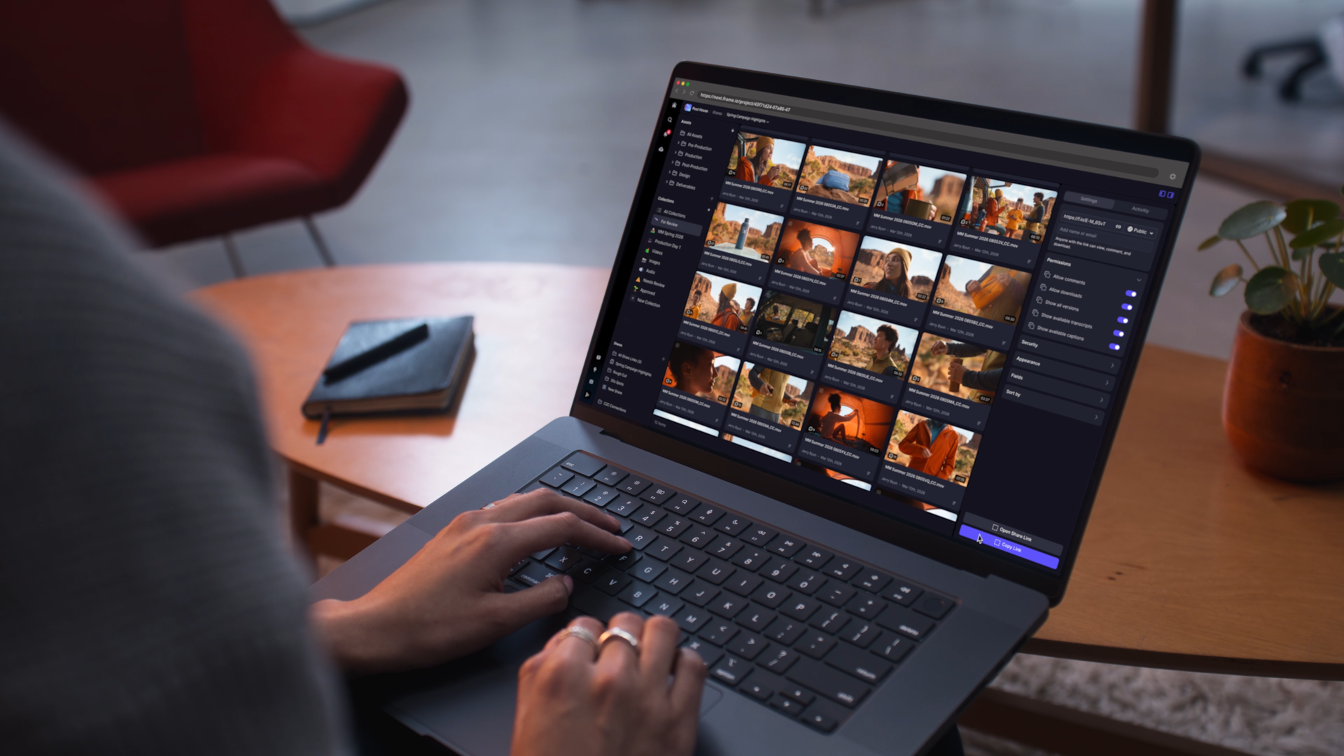 A person using a laptop with a dark photo management or photo editing software open. The screen displays thumbnail images of outdoor scenes with red rock formations, women in yellow jackets, and a sunset. The person's hand is on the keyboard, and they are wearing rings. The setting includes a wooden table, a planner or notebook with a pen, a red chair, and a potted plant nearby.