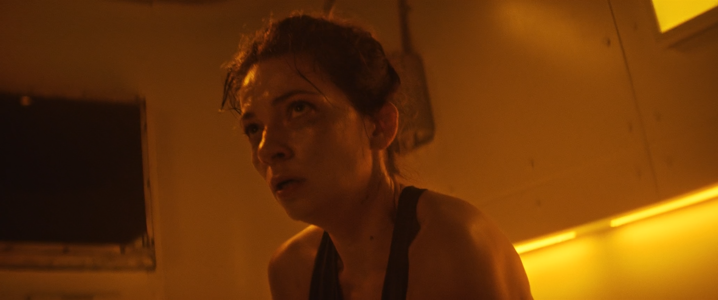 A woman with wet hair and a serious expression, illuminated by warm yellow lighting in an indoor setting.