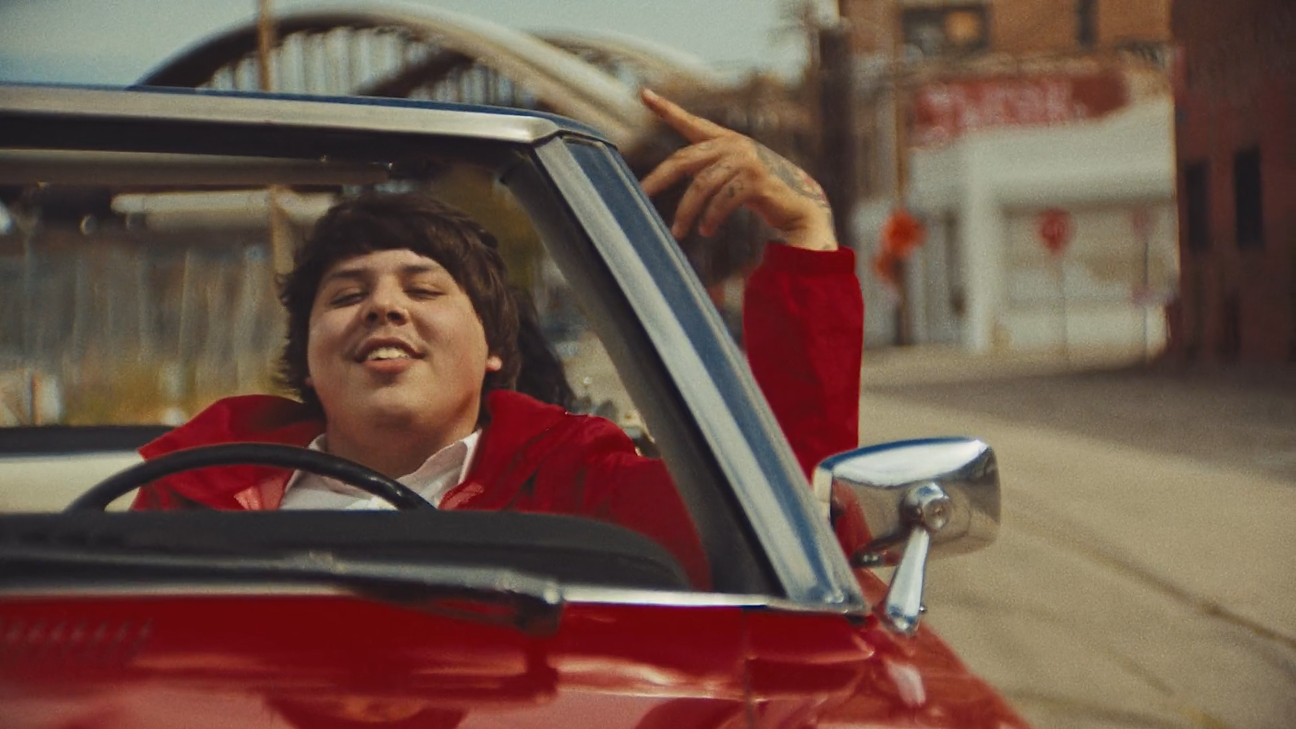 A young man with dark hair smiling and wearing a red jacket, sitting in the driver's seat of a red vintage convertible car, with his left hand raised and touching his head, on a street with buildings in the background.