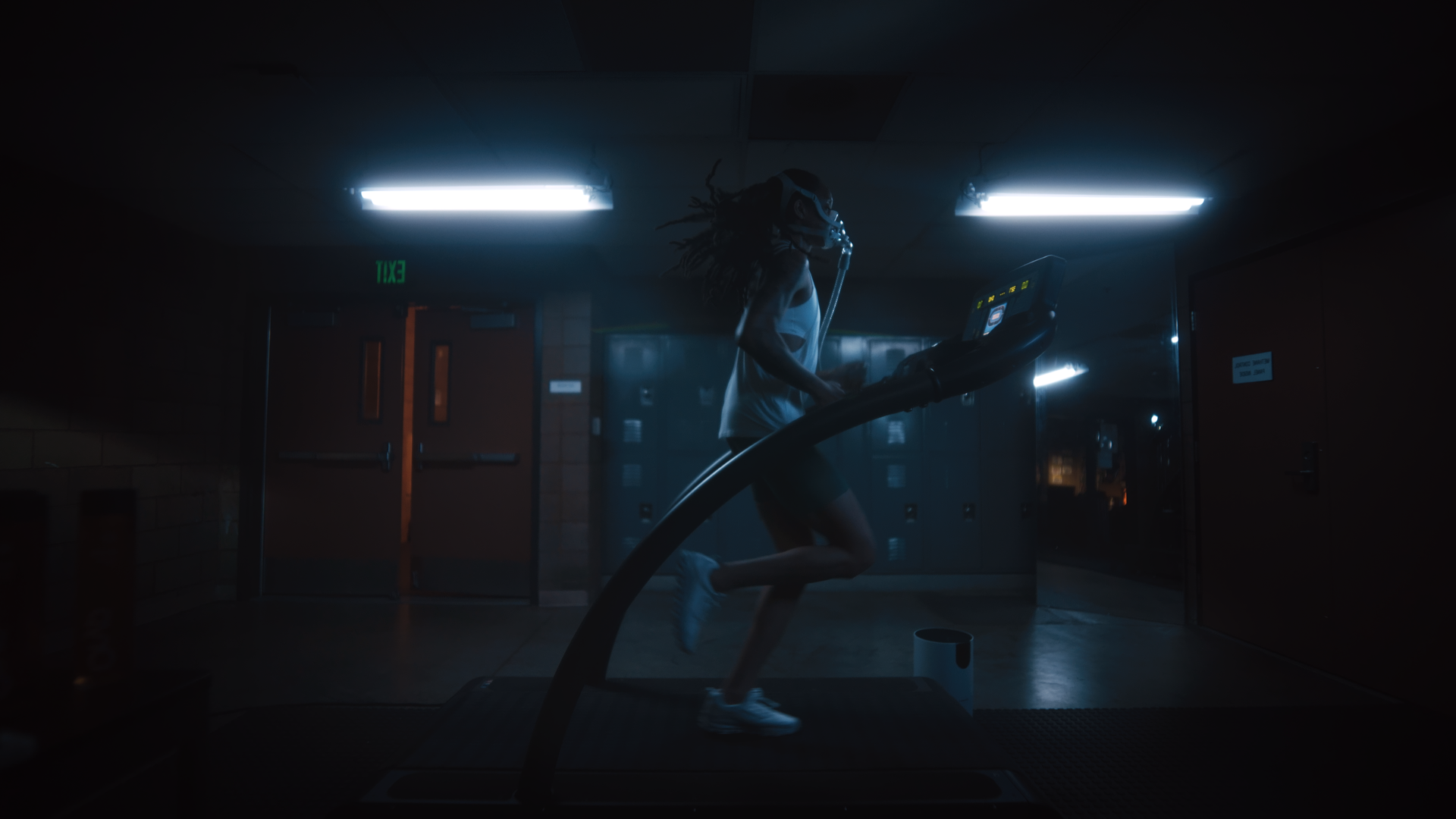 A person running on a treadmill in a dimly lit indoor gym, wearing a mask and headphones, with lockers and an exit sign in the background.
