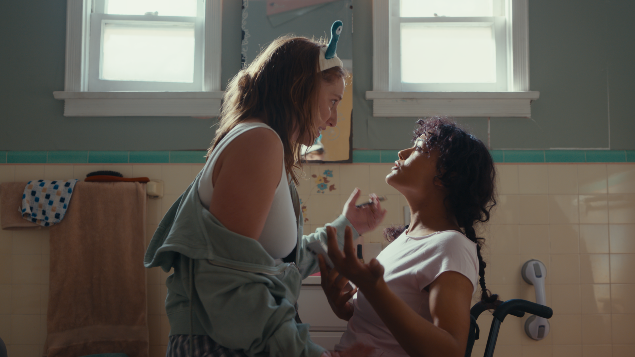 Two women, one standing and the other sitting, are in a bathroom with light from a window behind them. They are smiling and looking at each other, with one woman holding a small object and the other reaching for it. The woman standing has a headband with a plush toy on it.