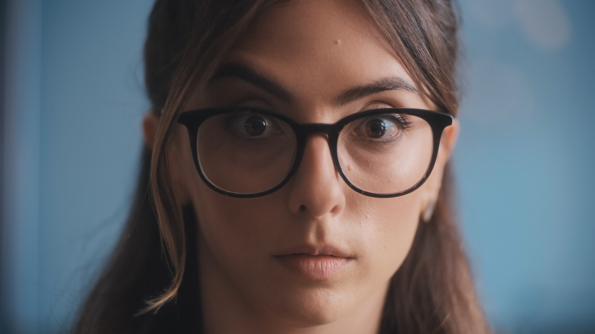 A close-up of a young woman with brown hair and glasses looking directly at the camera against a light blue background.