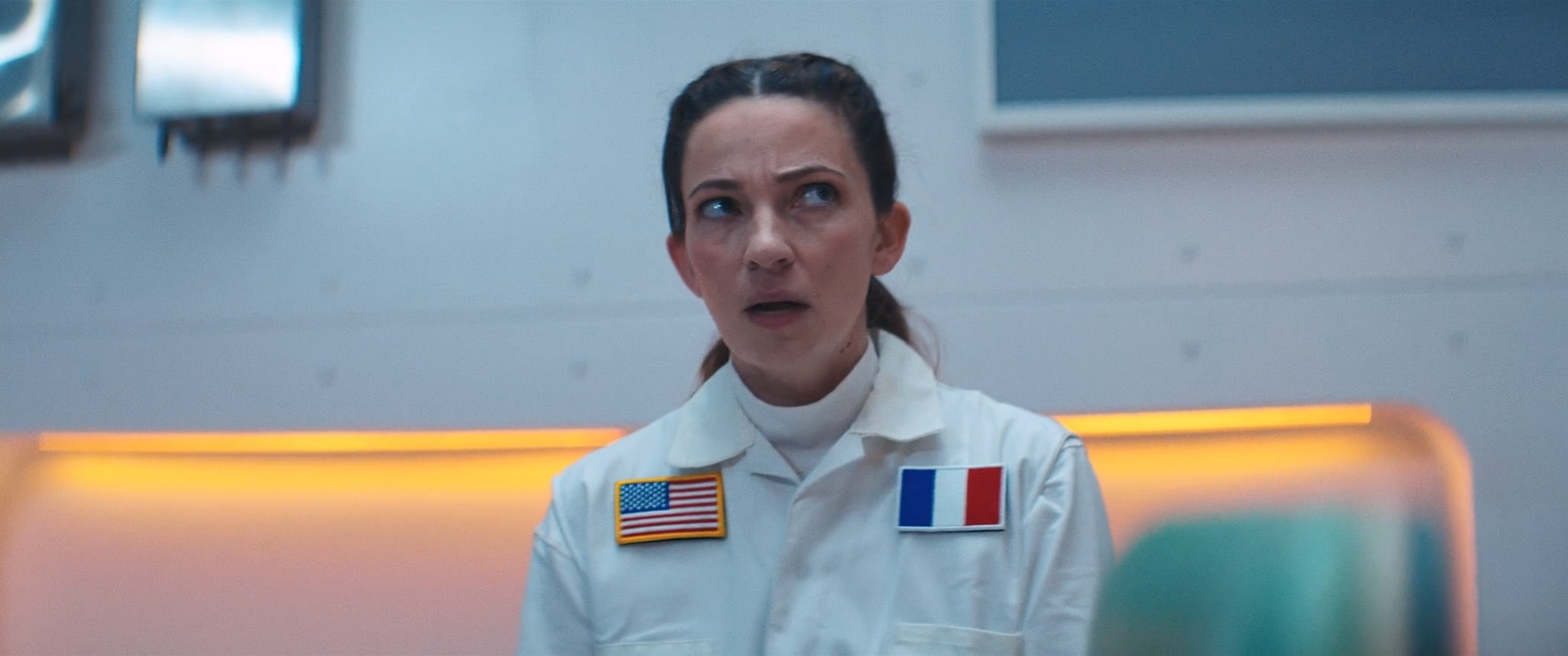 A woman dressed as an astronaut with French and American flags on her uniform, appearing surprised or confused in a sci-fi setting.