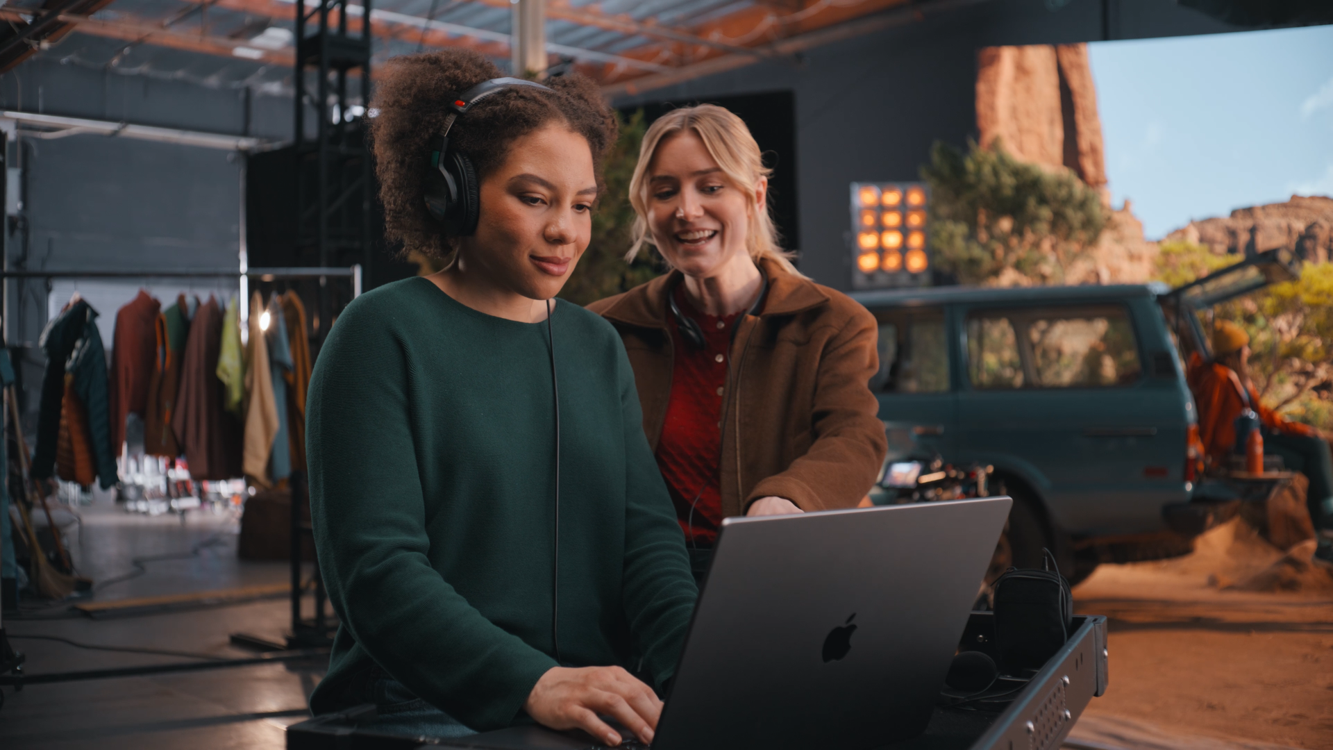 Two women working on a laptop at an outdoor filming set with vintage clothing and a vehicle in the background.