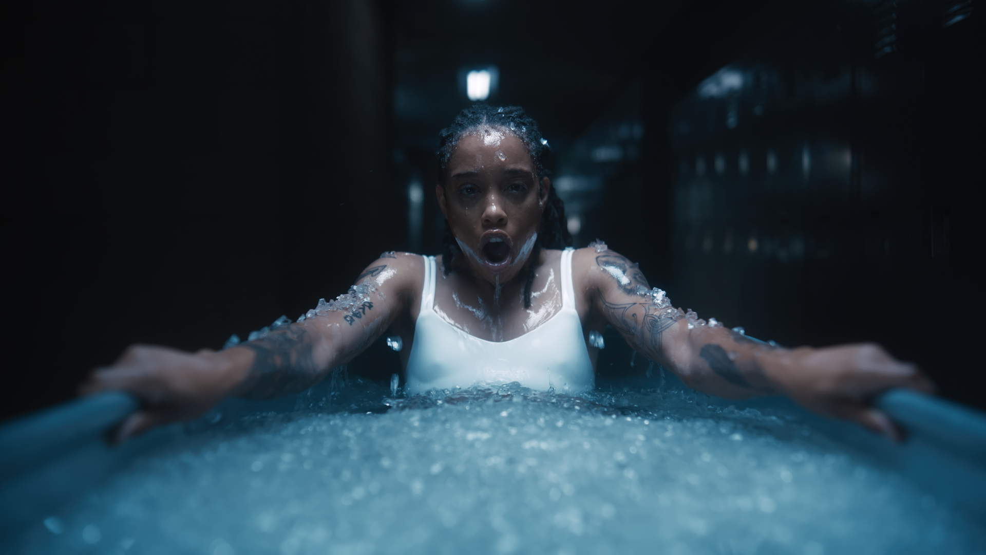A woman with tattoos on her arms and chest is in a swimming pool, with her arms outstretched and mouth open, appearing to be in a moment of effort or emotion.