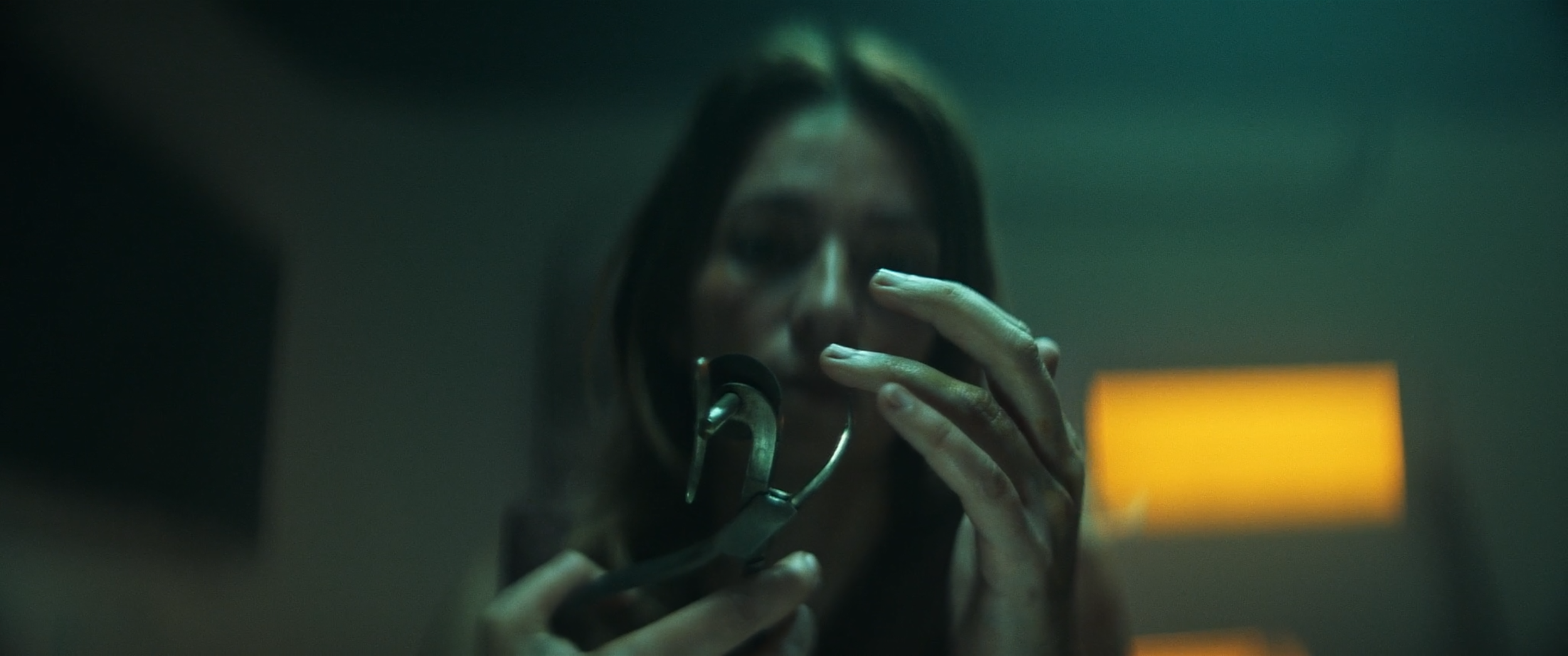 A woman with dark hair holds a phone to her face in a dimly lit room, wearing headphones around her neck, and appears to be talking into the phone.