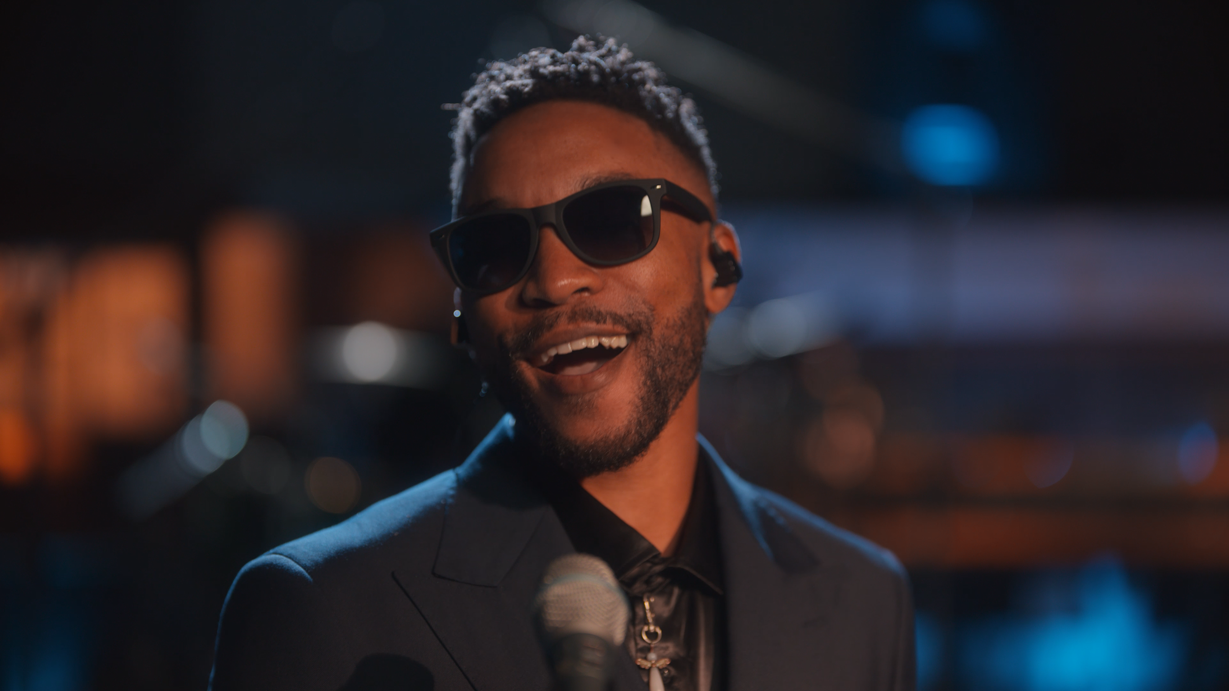 A man wearing sunglasses and a dark suit smiling at night with city lights in the background.