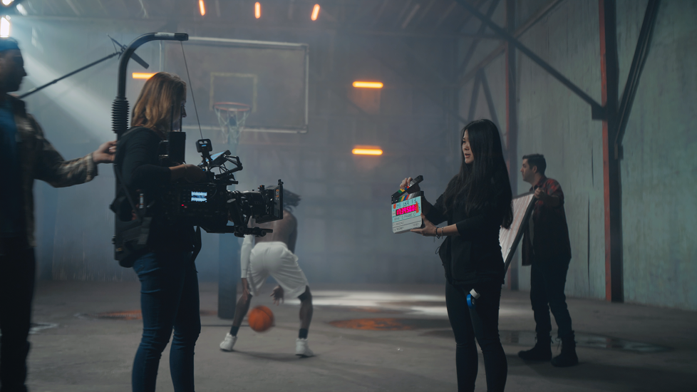 Behind the scenes of a film or video shoot in a gym, with crew and a woman holding a clapperboard, and a person playing basketball in the background.