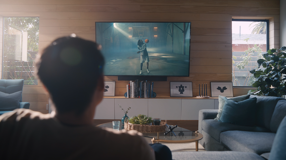 Person sitting on a gray couch watching a basketball game on a large flat-screen TV in a modern living room with wooden walls and large windows.