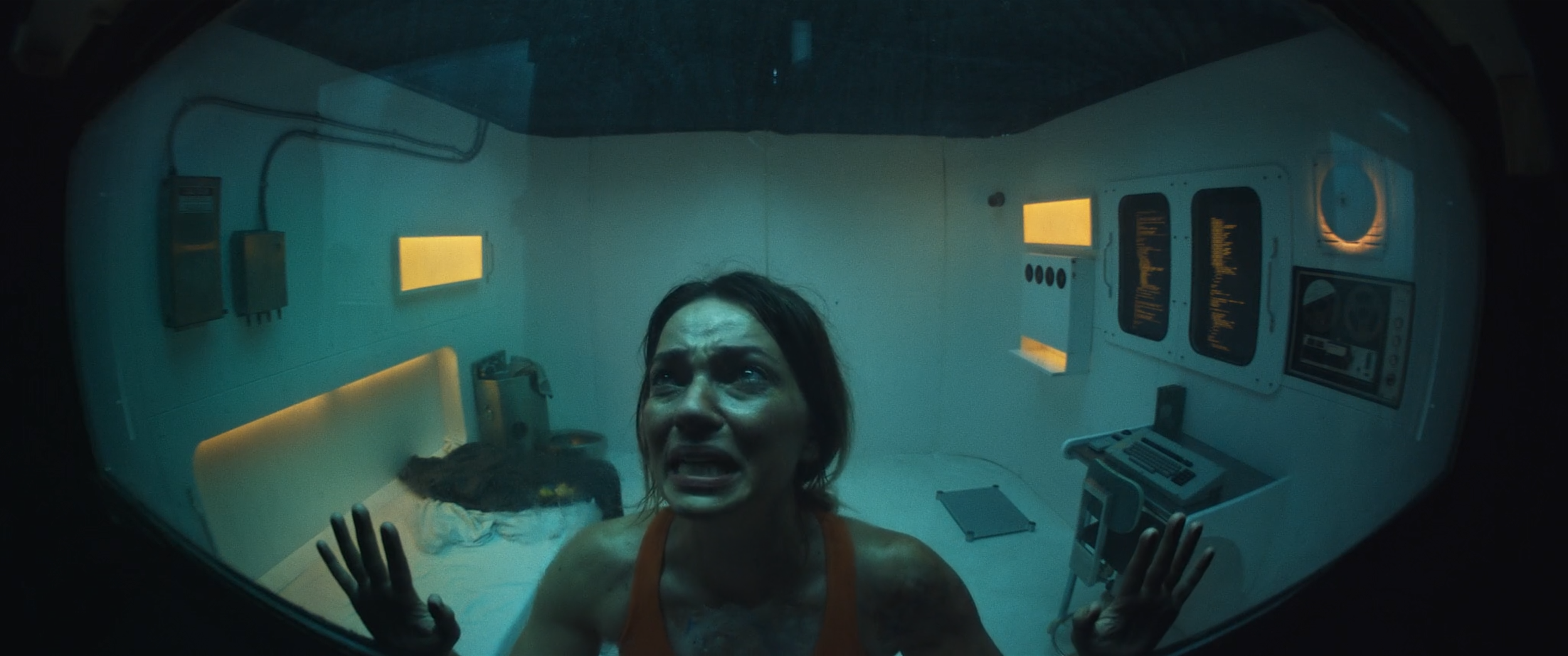 A woman with dark hair, wearing an orange tank top, is pressing her hands against a glass window and appears distressed inside a medical or scientific chamber with monitors and equipment on the walls.
