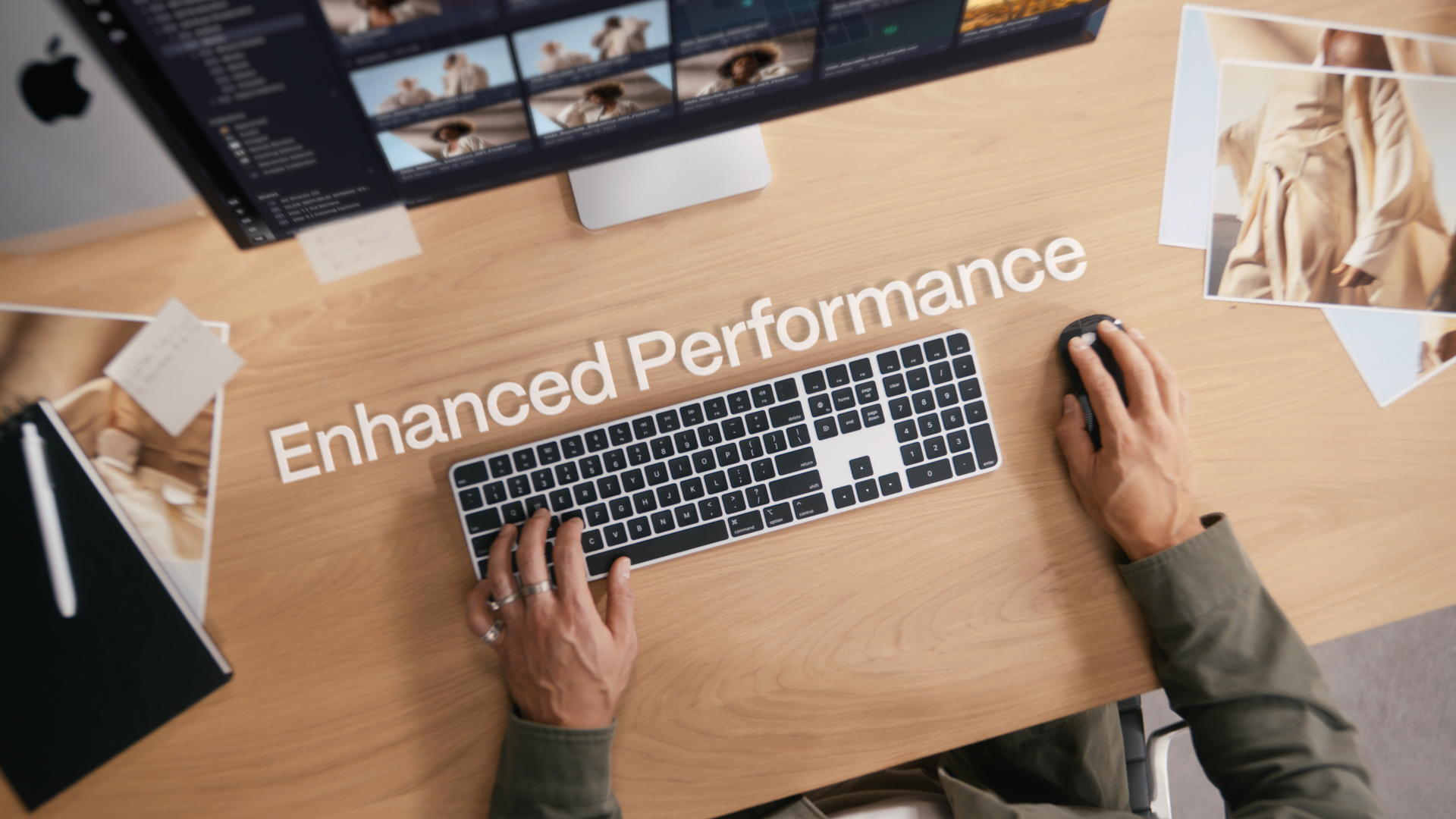Person using a computer mouse and keyboard on a wooden desk with the words 'Enhanced Performance' written on the desk. There are printed photographs and a monitor displaying a photo editing or video editing software.
