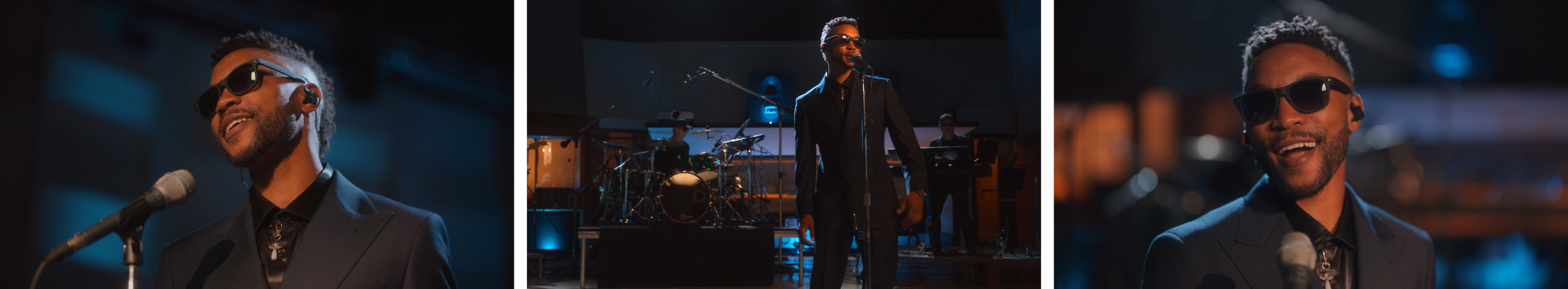A man wearing dark sunglasses and a black suit singing into a microphone during a live performance, with a band playing in the background.