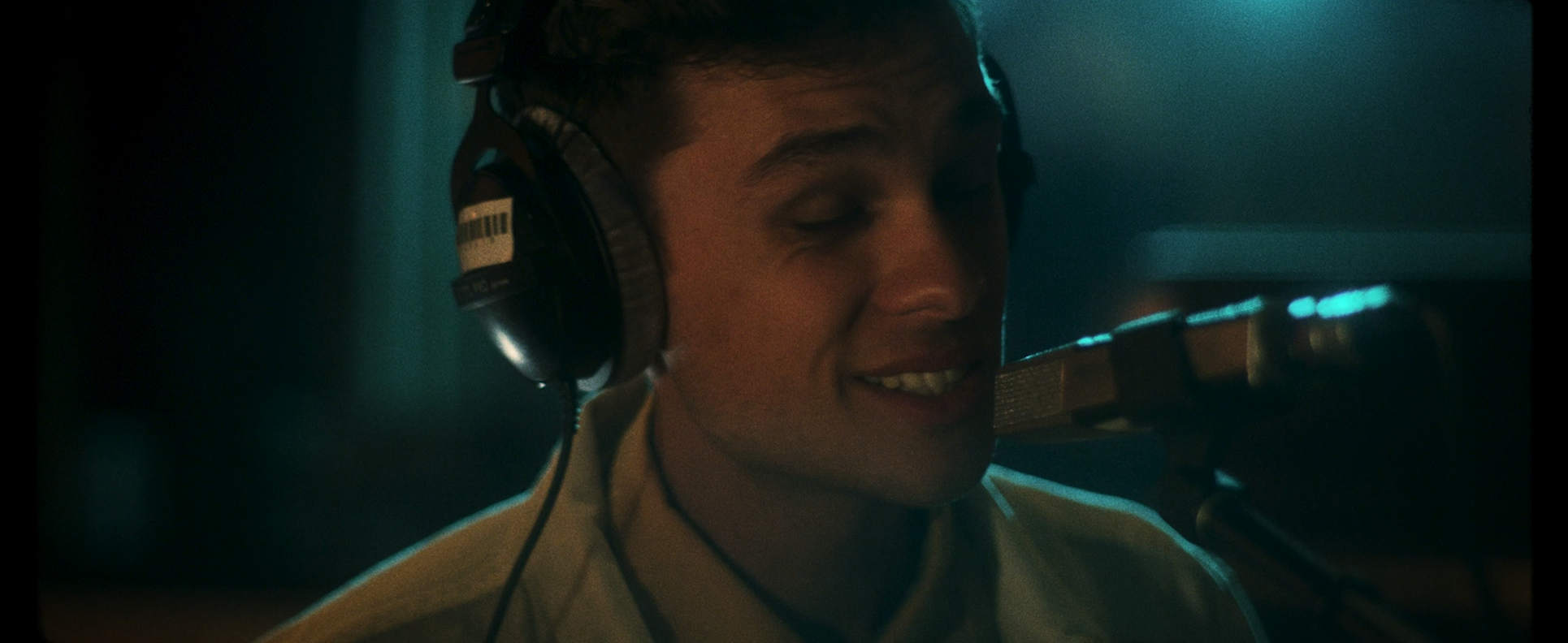 A young man wearing headphones is singing into a microphone in a recording studio with his eyes closed and a relaxed expression.