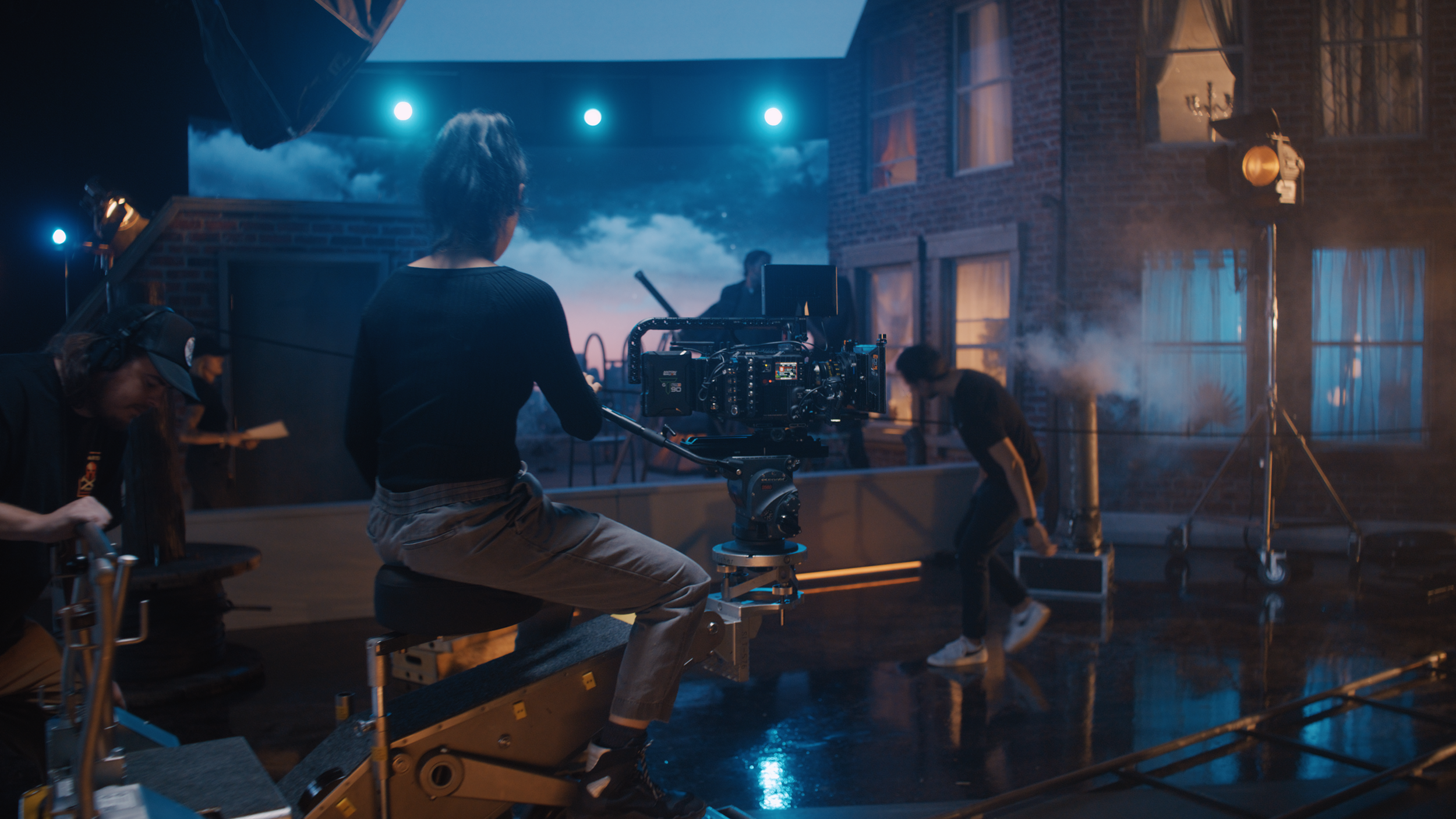 Film crew setting up a shot outdoors at night with a brick building, smoke, and blue lighting.