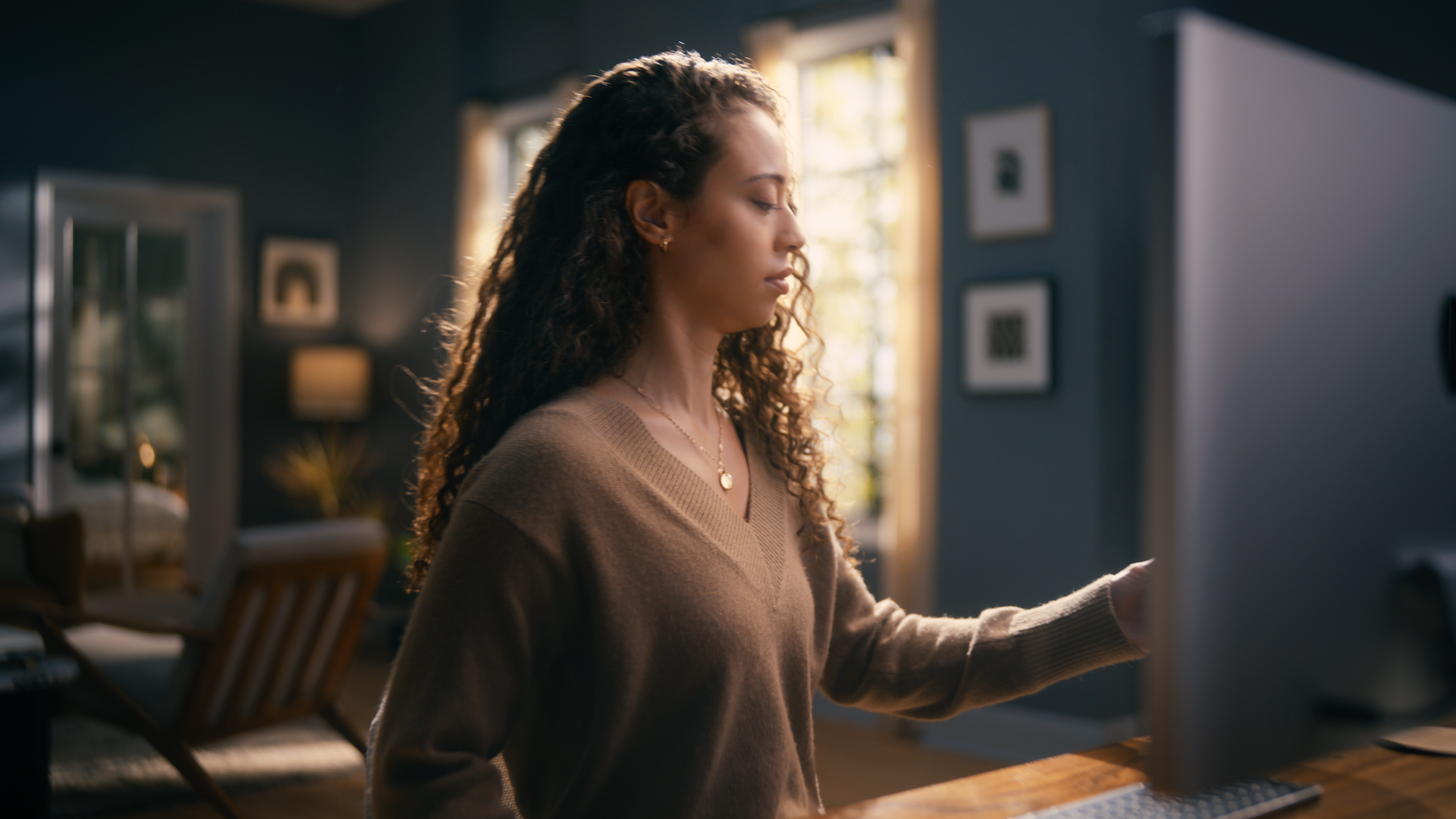 A woman with curly hair in a brown sweater looking at a computer screen in a cozy, well-lit living room.
