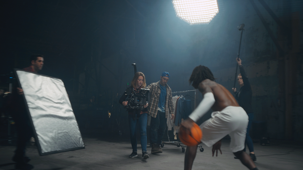 Behind the scenes of a photoshoot with a model in basketball attire and crew members adjusting equipment in an industrial-style studio.