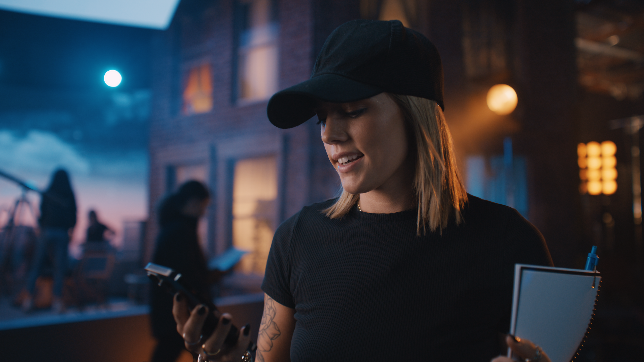 Young woman with blonde hair wearing a black baseball cap and black shirt, looking at her phone in an outdoor setting during dusk, with people and a brick building in the background.
