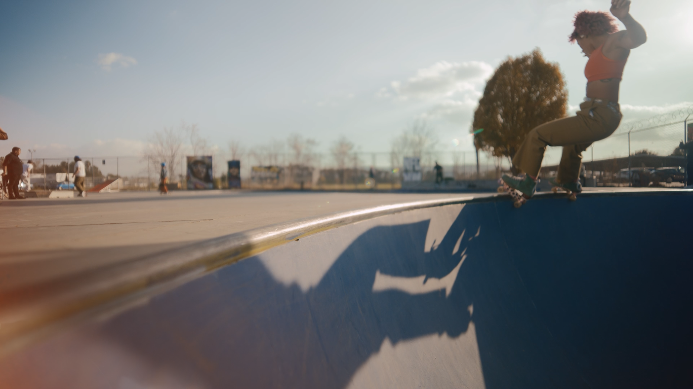 A person with curly hair skateboarding on the edge of a skateboard bowl at a skate park on a sunny day. Other people are visible in the background.