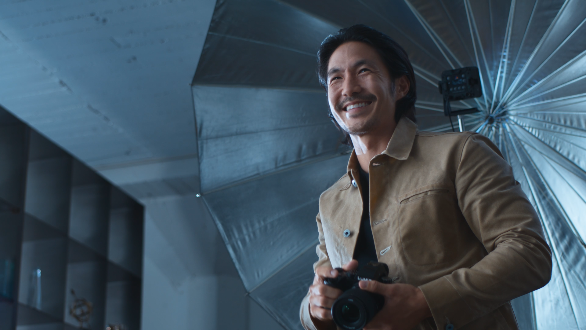 A man smiling while holding a camera in front of a photography studio with large umbrella lights and reflective surfaces.