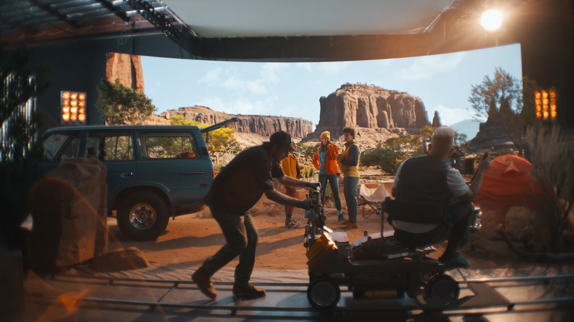Filmmaking crew filming a scene in a desert landscape with large rock formations; crew operating camera and equipment, people standing and talking, a blue van parked nearby, bright sunlight