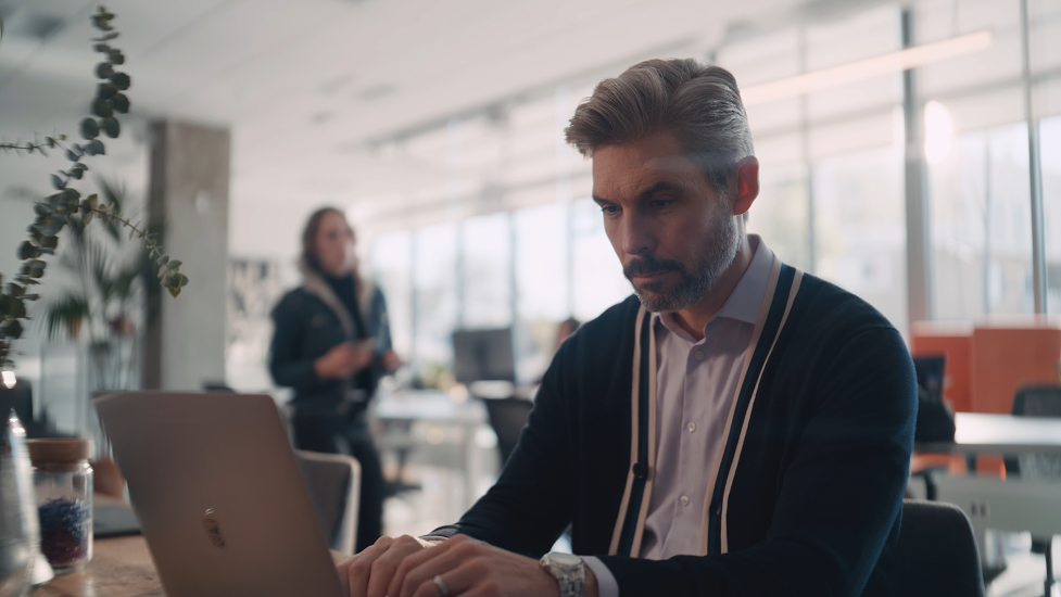 A man working on a laptop in a bright, modern office with a woman in the background holding a phone.