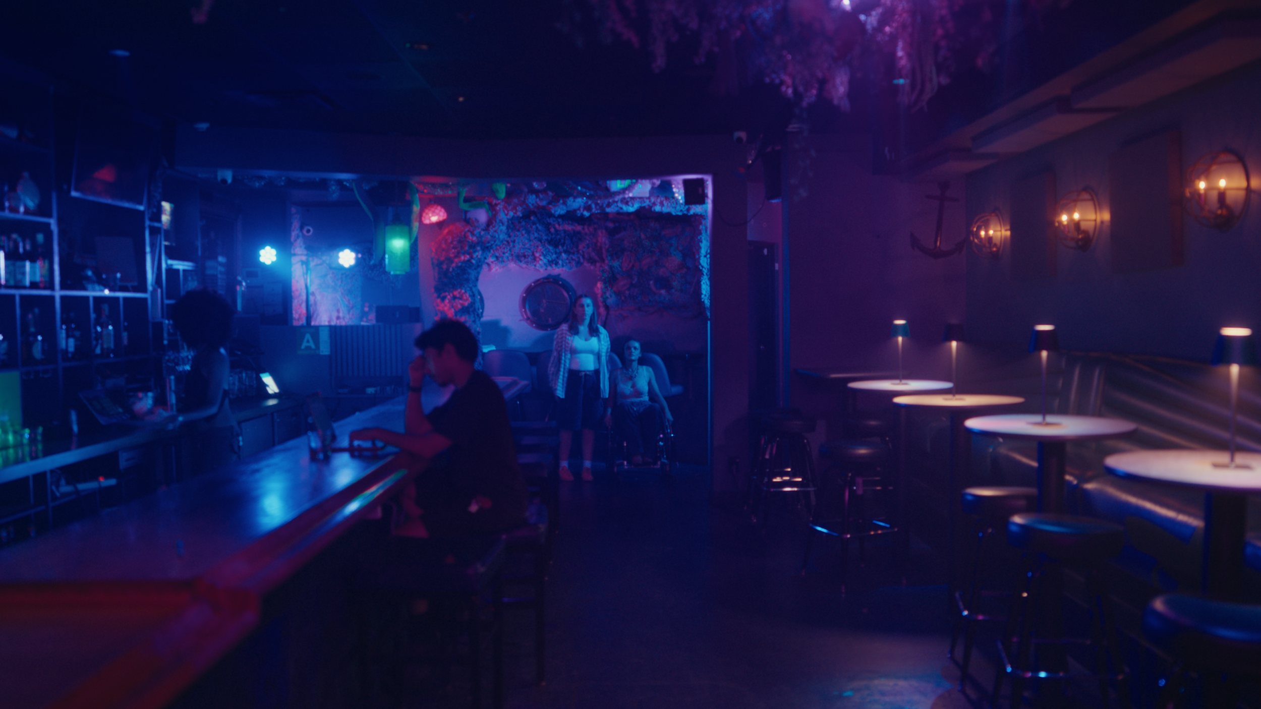 A dimly lit nightclub with a bar area, a few patrons, and blue and purple lighting. Two women are sitting and standing near the stage, which has colorful decorations.