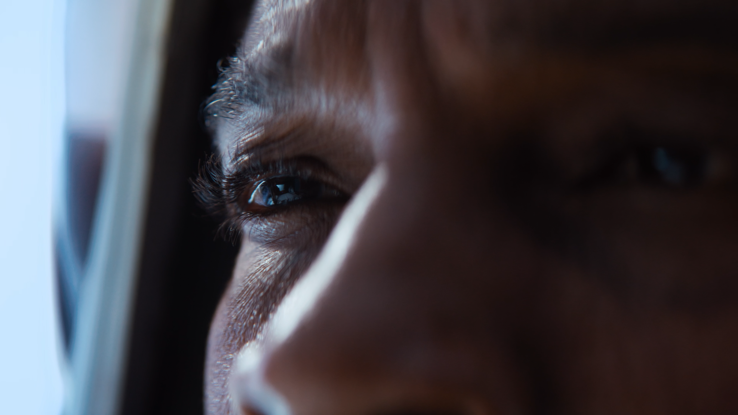 Close-up of a person's face as they look out of a window, with focus on their eye and forehead, showing dark eyelashes and eyebrow hairs.