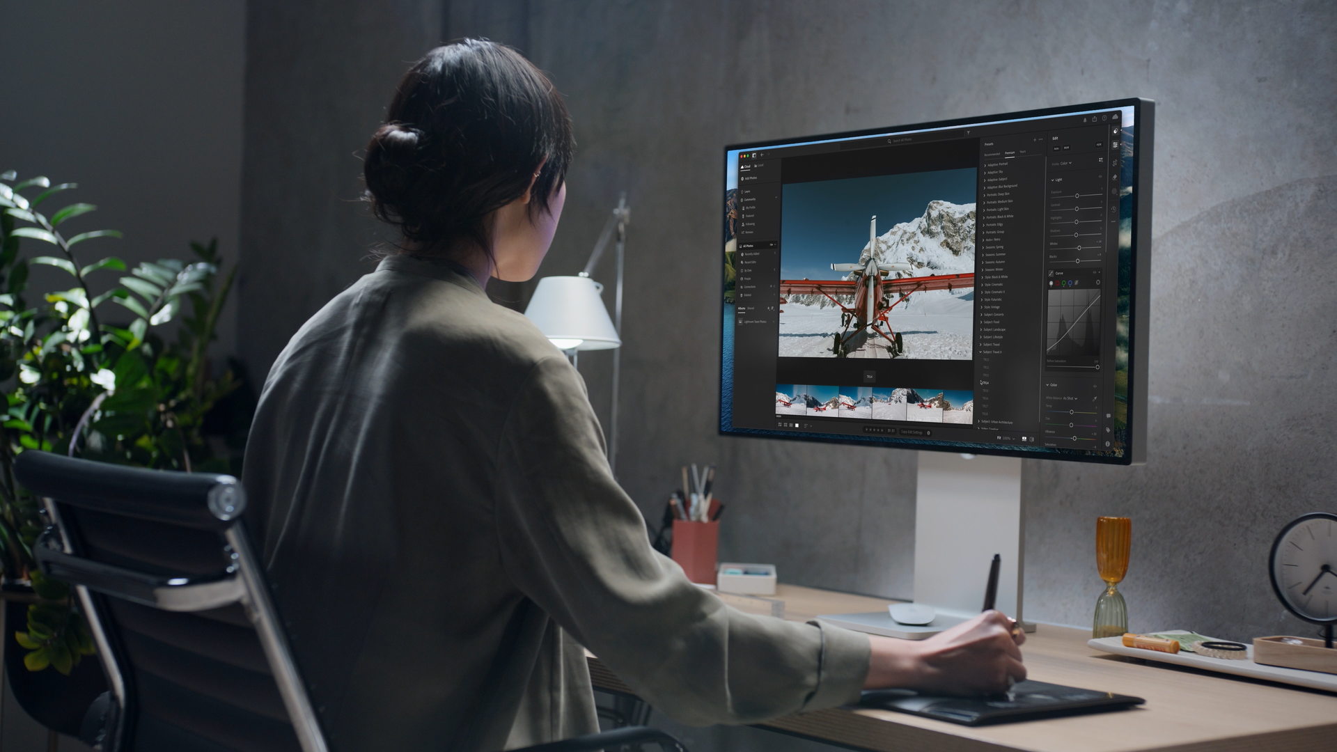 A woman sitting at a desk editing a photo of an airplane in a snowy mountain landscape on a large monitor, surrounded by plants and office supplies.
