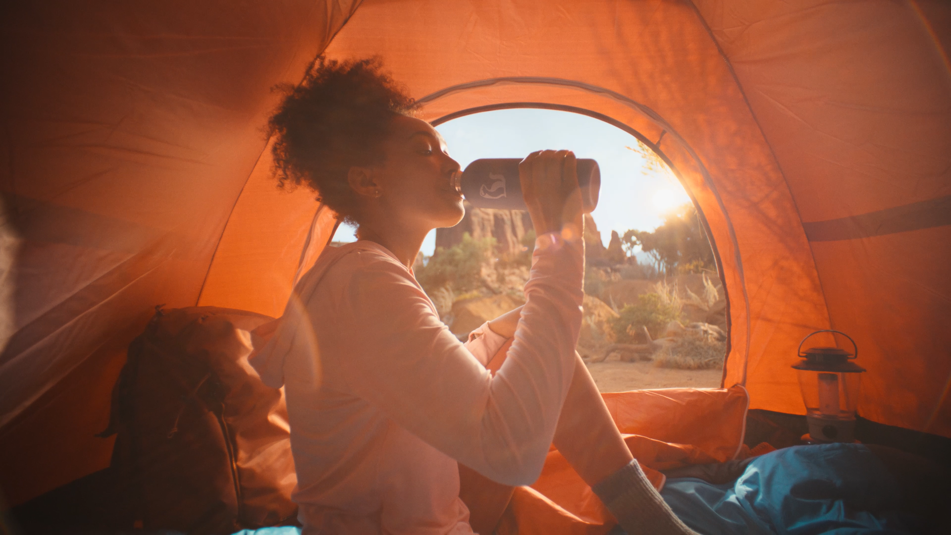 A woman with curly hair drinking from a tumbler inside an orange camping tent at sunset, with outdoor scenery of rocks and trees visible through the tent's opening.
