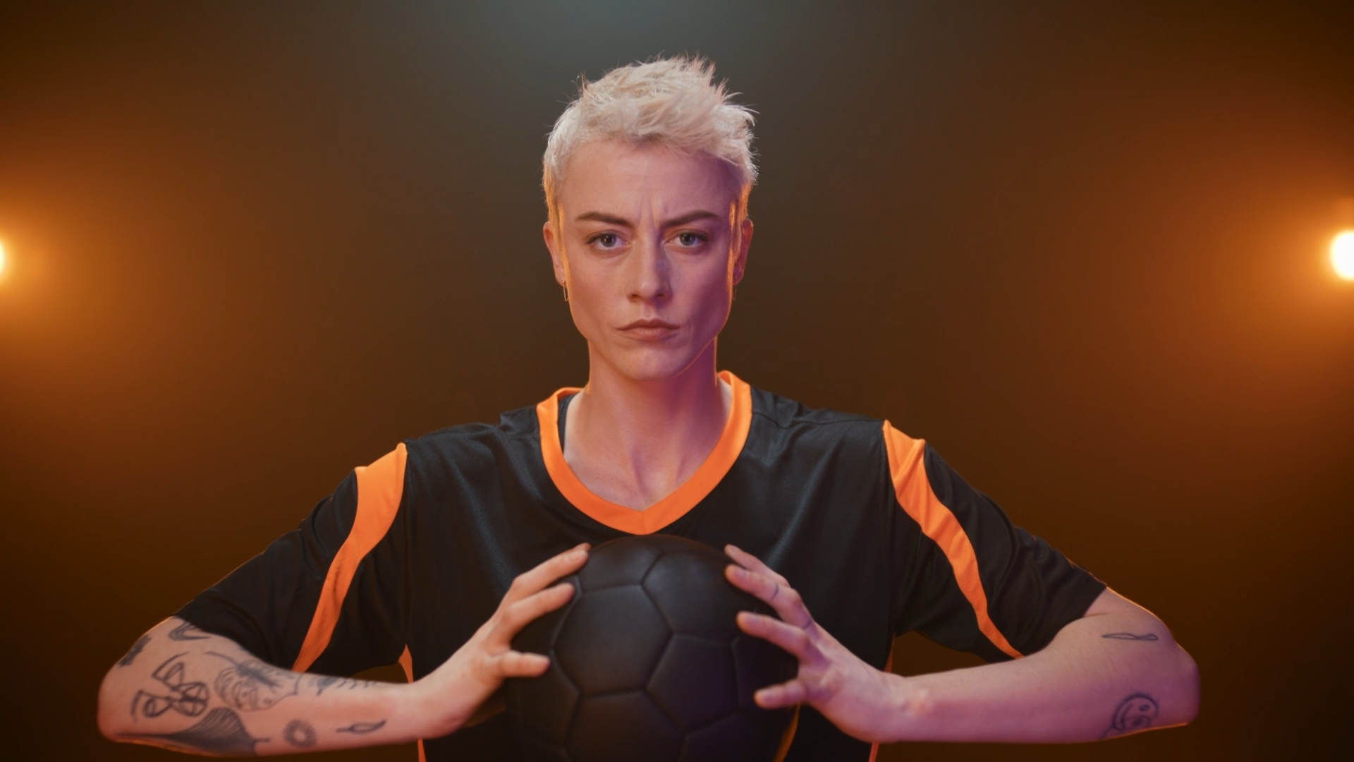 A woman with short blonde hair in a black and orange sports jersey holding a black soccer ball, standing with a serious expression in front of a dark background with warm lights.