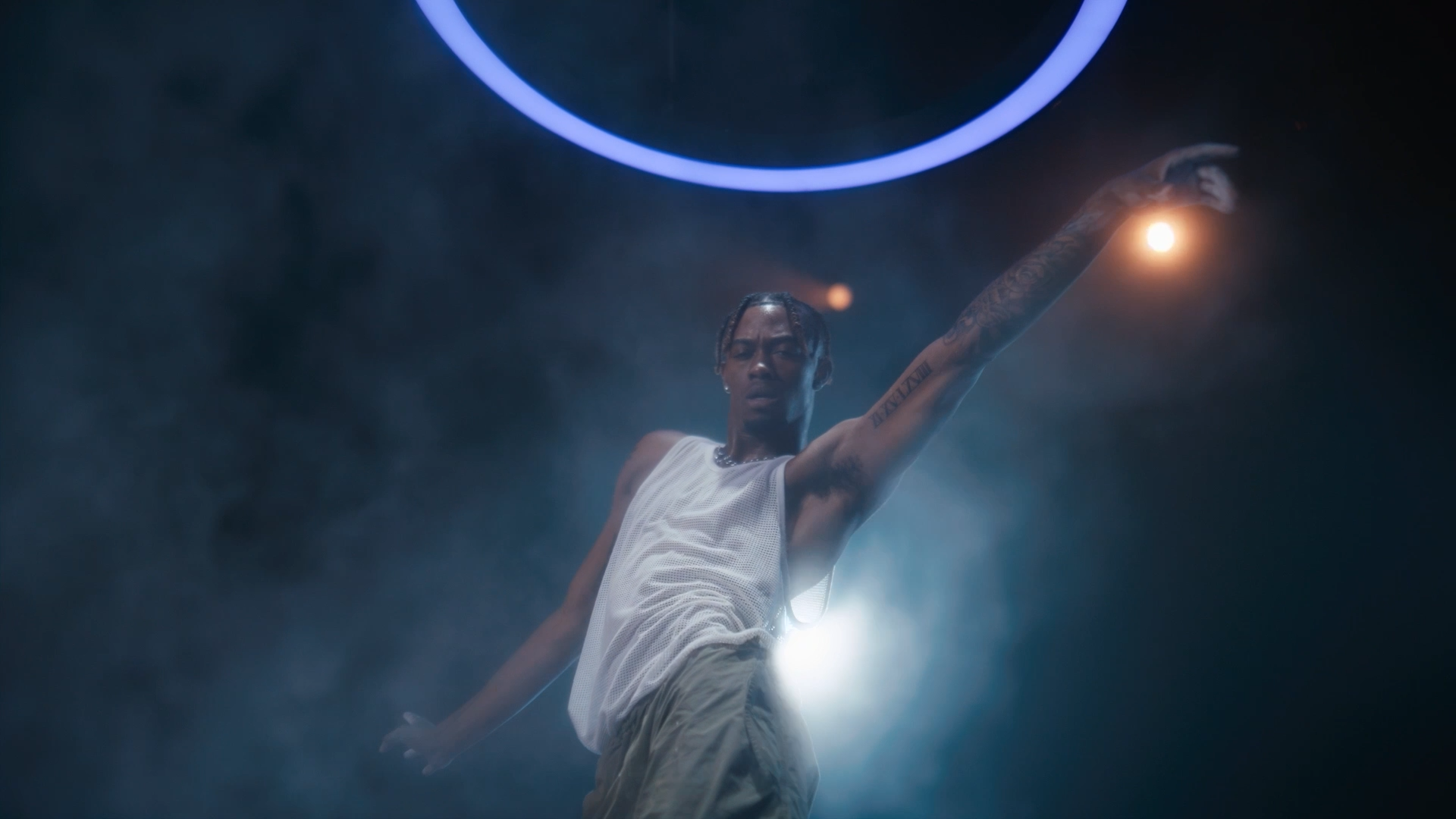 A young man with tattoos on his arm is dancing on stage in front of bright lights, wearing a sleeveless white shirt and beige pants.