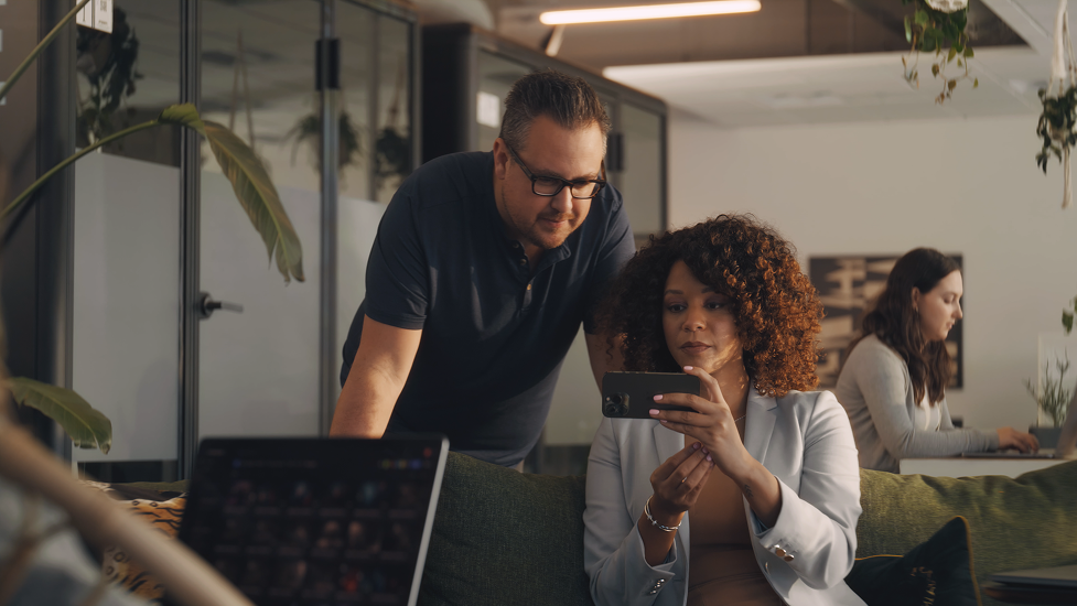 Two people working together in an office, with a woman looking at her phone and a man leaning over to see the screen, while another woman works at her desk in the background.