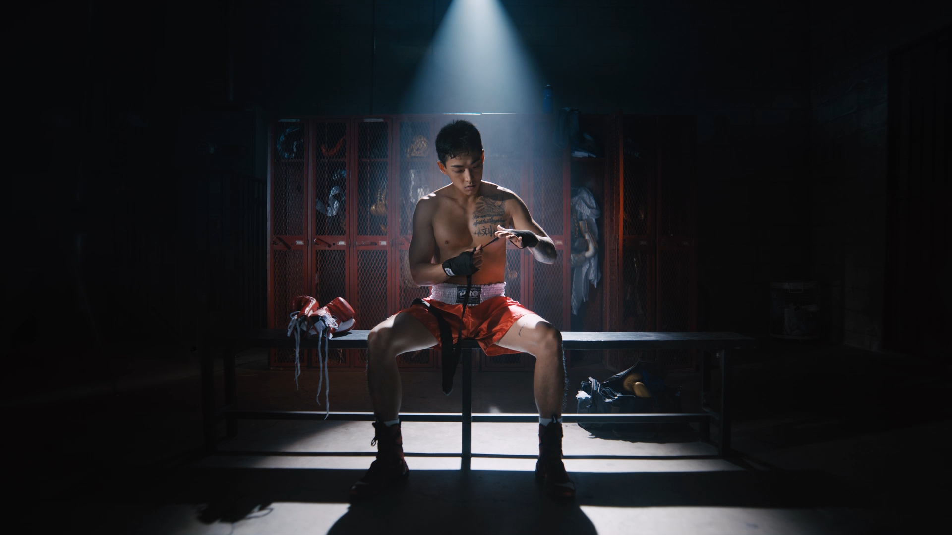 A male boxer with tattoos and wearing red shorts and black training gloves sits on a bench in a dimly lit locker room, preparing for a fight by wrapping his hands.