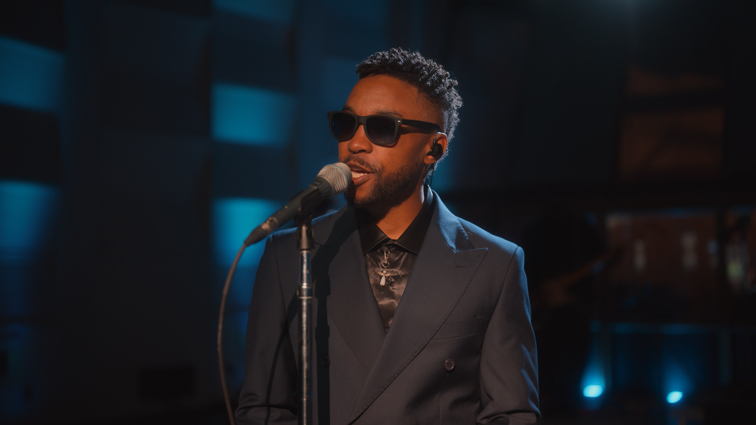 A man wearing dark sunglasses and a dark suit singing into a microphone in a recording studio.