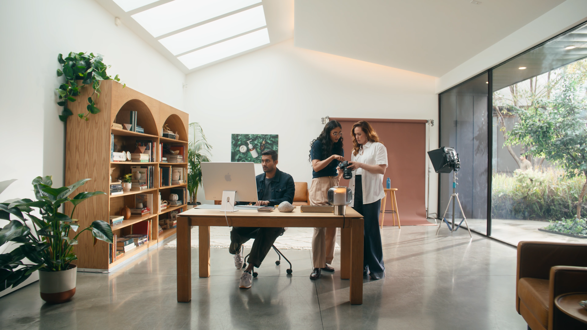 Three people in a modern, bright photo studio during a photography session. A woman holds a camera, and another woman shows her photos, while a man sits at a desk with a computer. Studio equipment includes a large near-floor light, a backdrop, and a large window with a garden view. There are books and plants on a wooden bookshelf along the wall.
