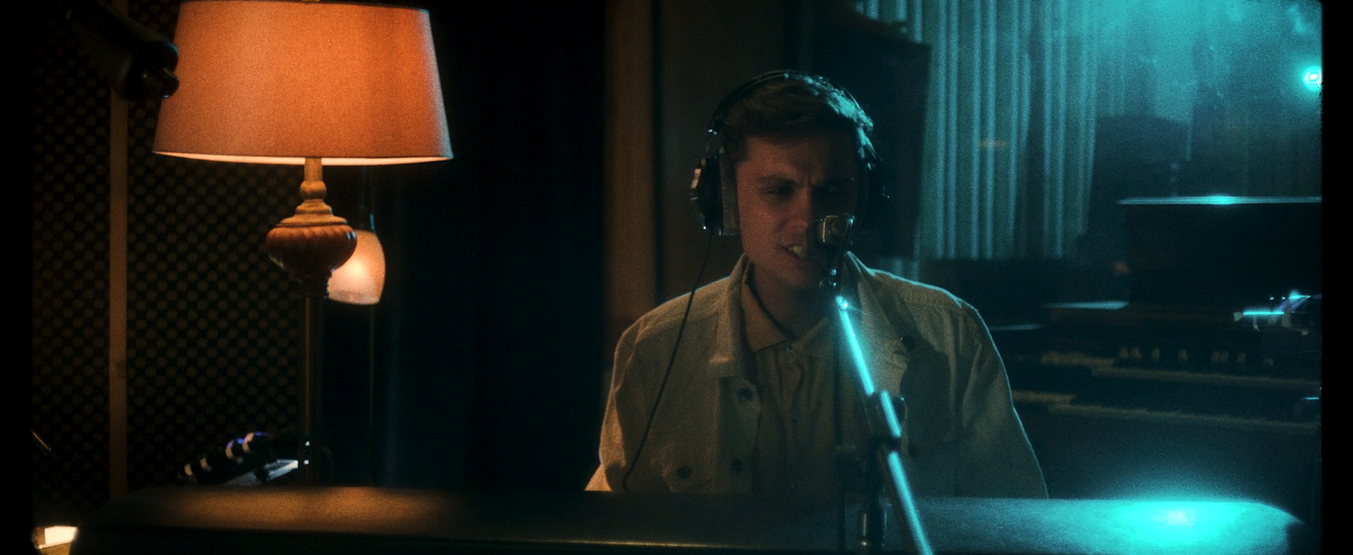 A young man wearing headphones, singing or speaking into a microphone in a recording studio with dim lighting, a lamp, and a piano in the background.