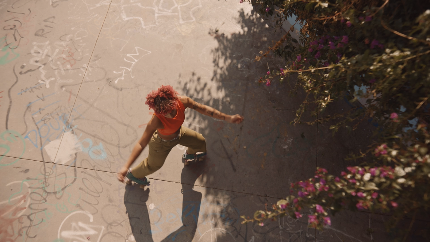 A woman with pink curly hair wearing an orange sleeveless top and beige pants stepping on a concrete sidewalk with colorful graffiti, near pink flowers on bushes.
