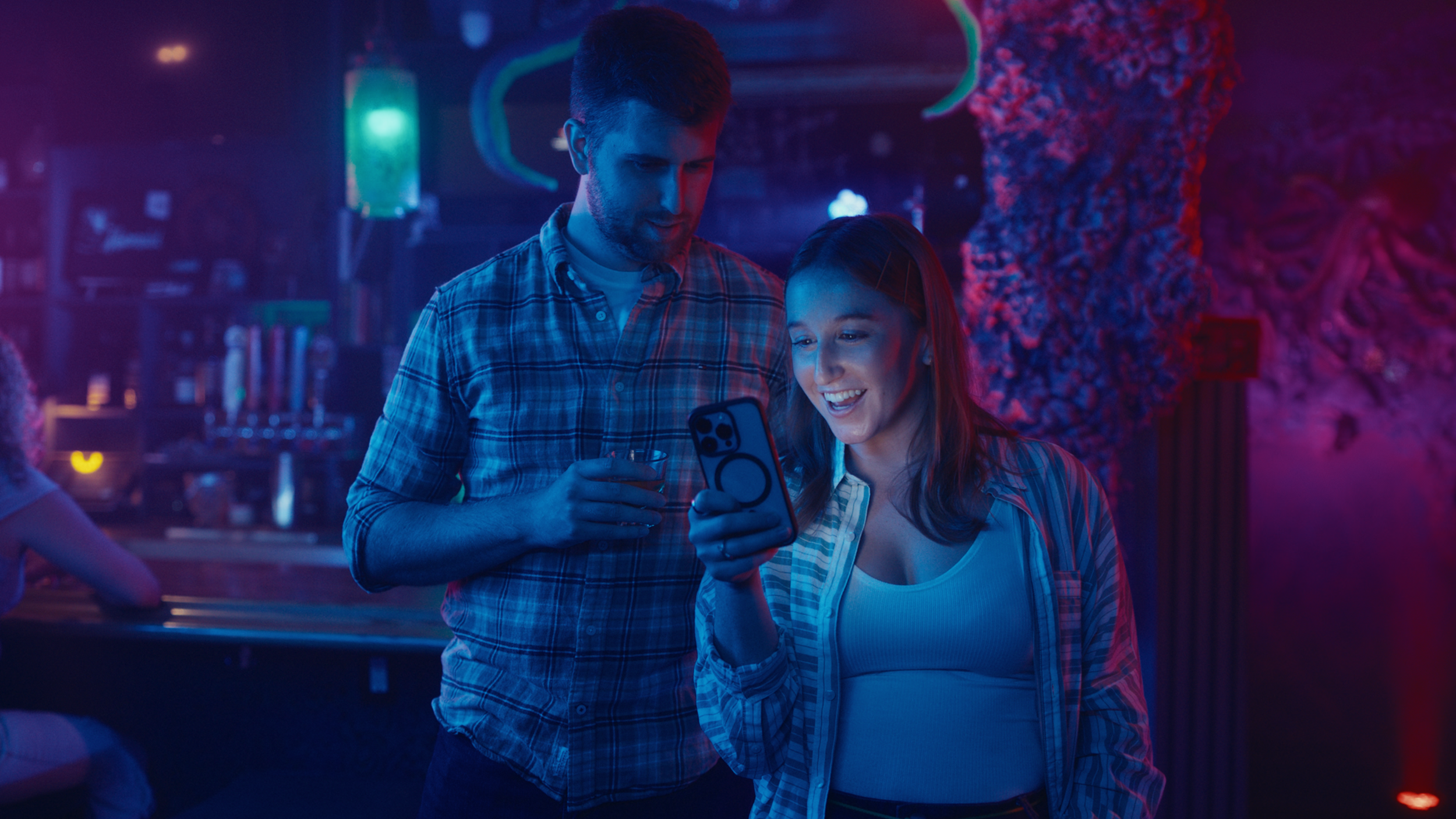 Two young adults, a man and a woman, are in a dimly lit bar or club with colorful neon lighting. They are looking at a smartphone together, smiling. The man holds a drink, and the woman appears excited or happy while viewing the phone.
