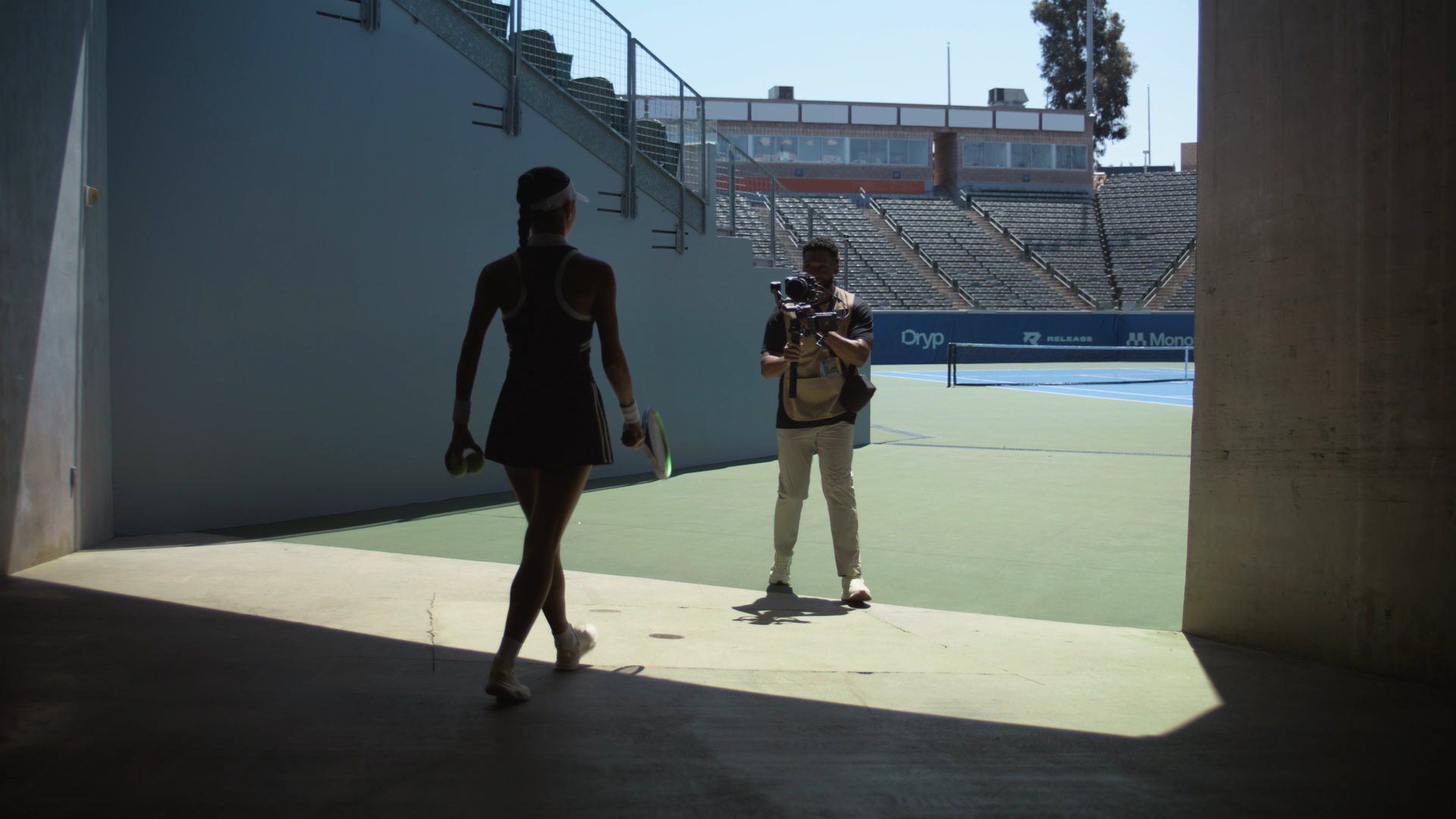 A female tennis player walking towards a male cameraman on a tennis court, with empty stadium seats in the background.