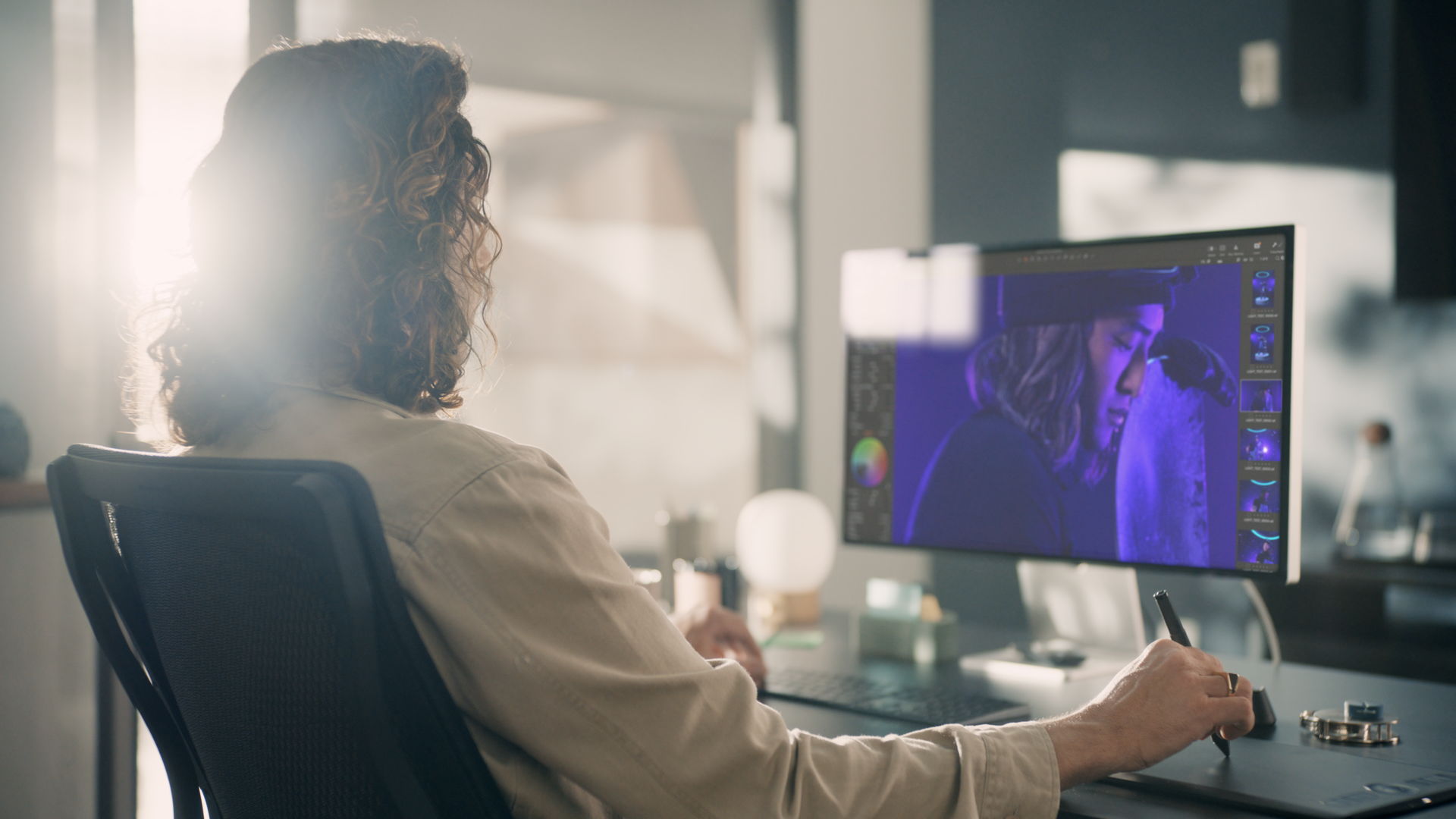 A woman with wavy hair working on digital photo editing at a desk in a modern, well-lit office, with her monitor displaying a portrait of a woman with purple lighting.