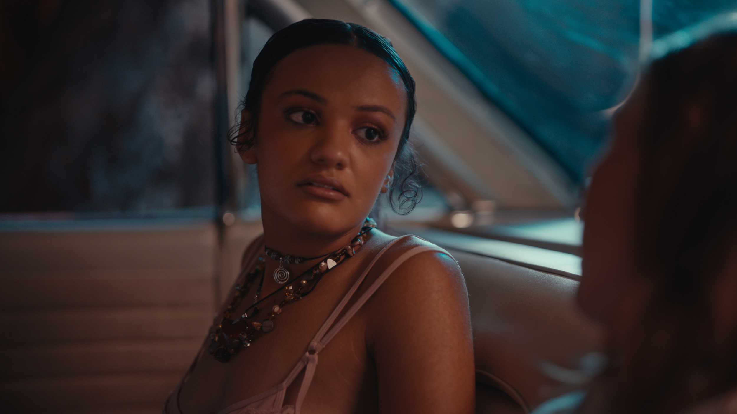 A woman with dark hair styled in baby hairs, wearing multiple necklaces, sitting inside a vehicle, looking at another person with a serious expression.