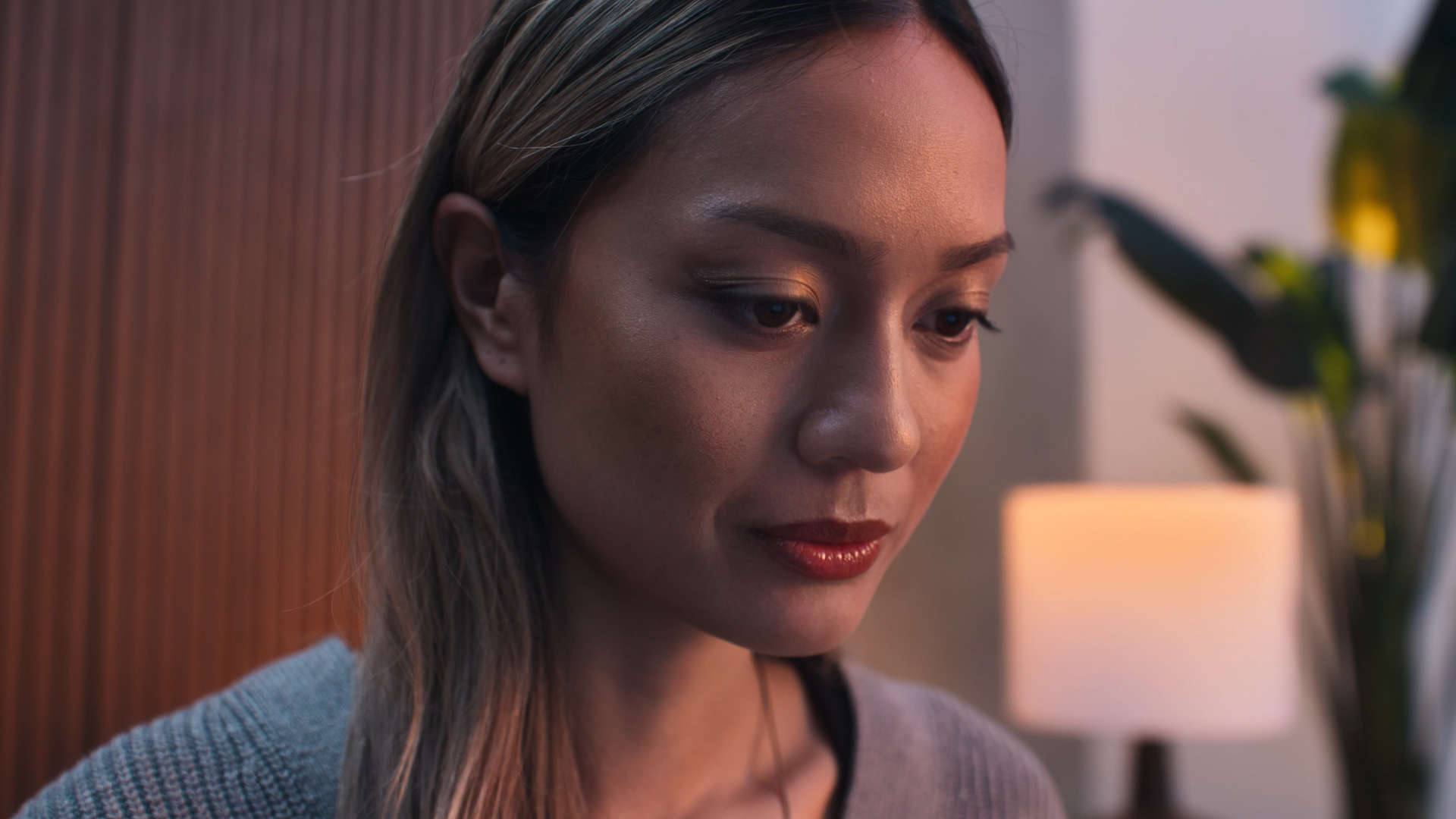 A young woman with light skin and long, light brown hair looking downward, standing indoors near a warm-colored lamp and a plant.