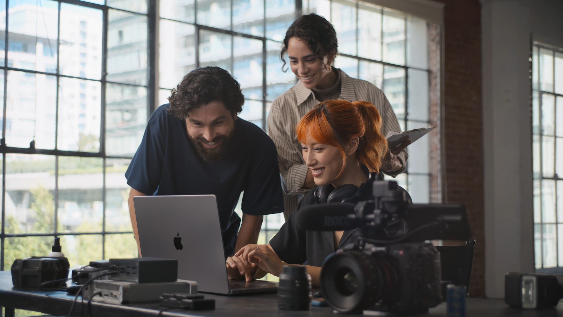 Four people gathered around a laptop in a well-lit industrial-style room, with camera equipment and electronic devices on the table.