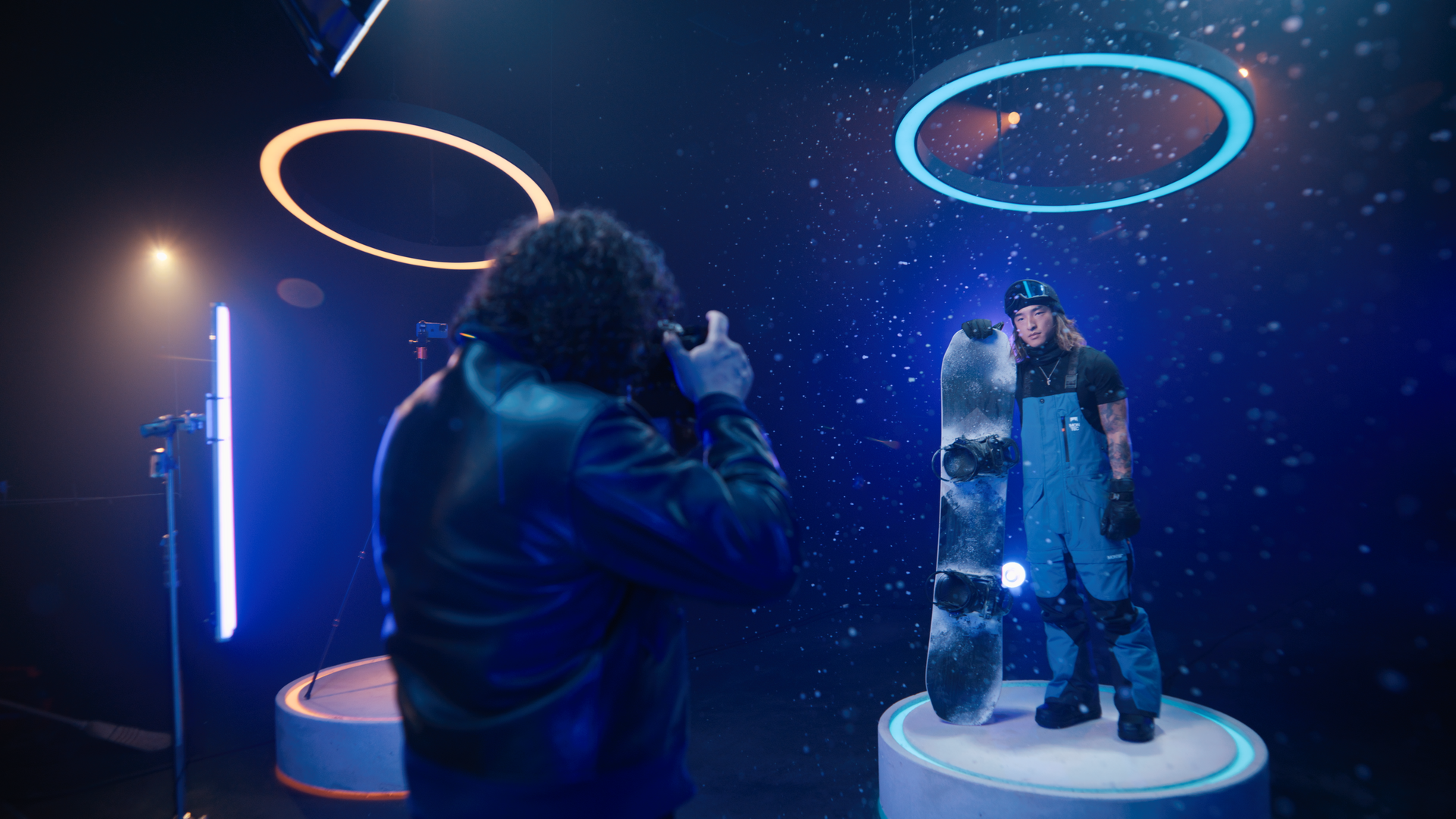 Person in snowboard gear posing with snowboard on display in a futuristic, illuminated setting with blue and orange ring lights and floating particles