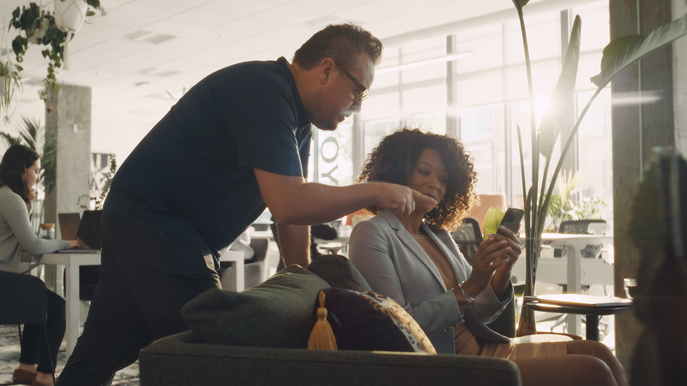 A man and a woman in a cafe, with the man leaning over and pointing at the woman's smartphone while she looks at it.