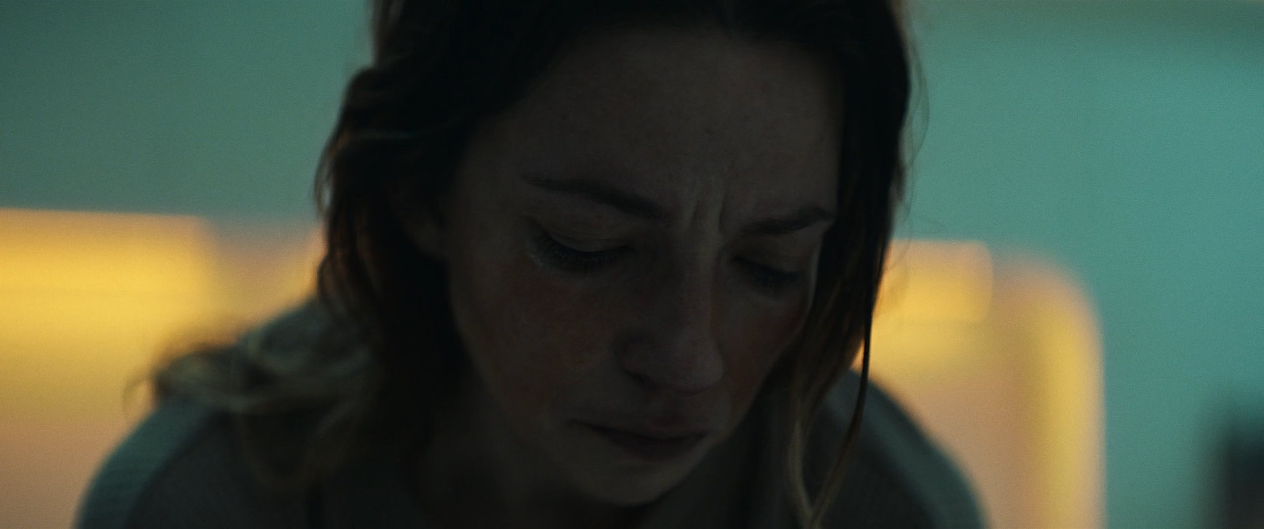 Close-up of a woman with wet hair and a concerned expression, looking down, with soft, colorful background lighting.