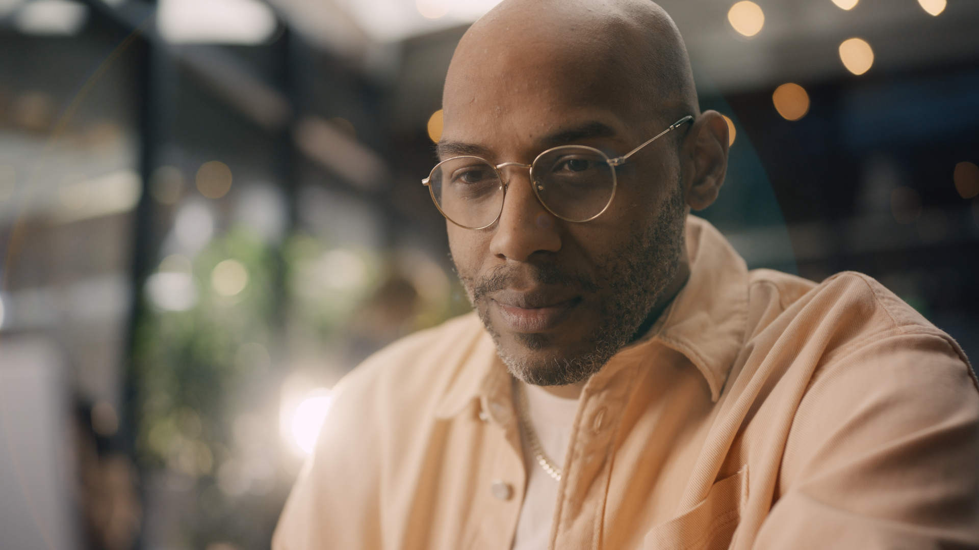 A man with a shaved head and glasses sitting at a table in a cafe, looking down thoughtfully, with blurred warm lighting and greenery in the background.
