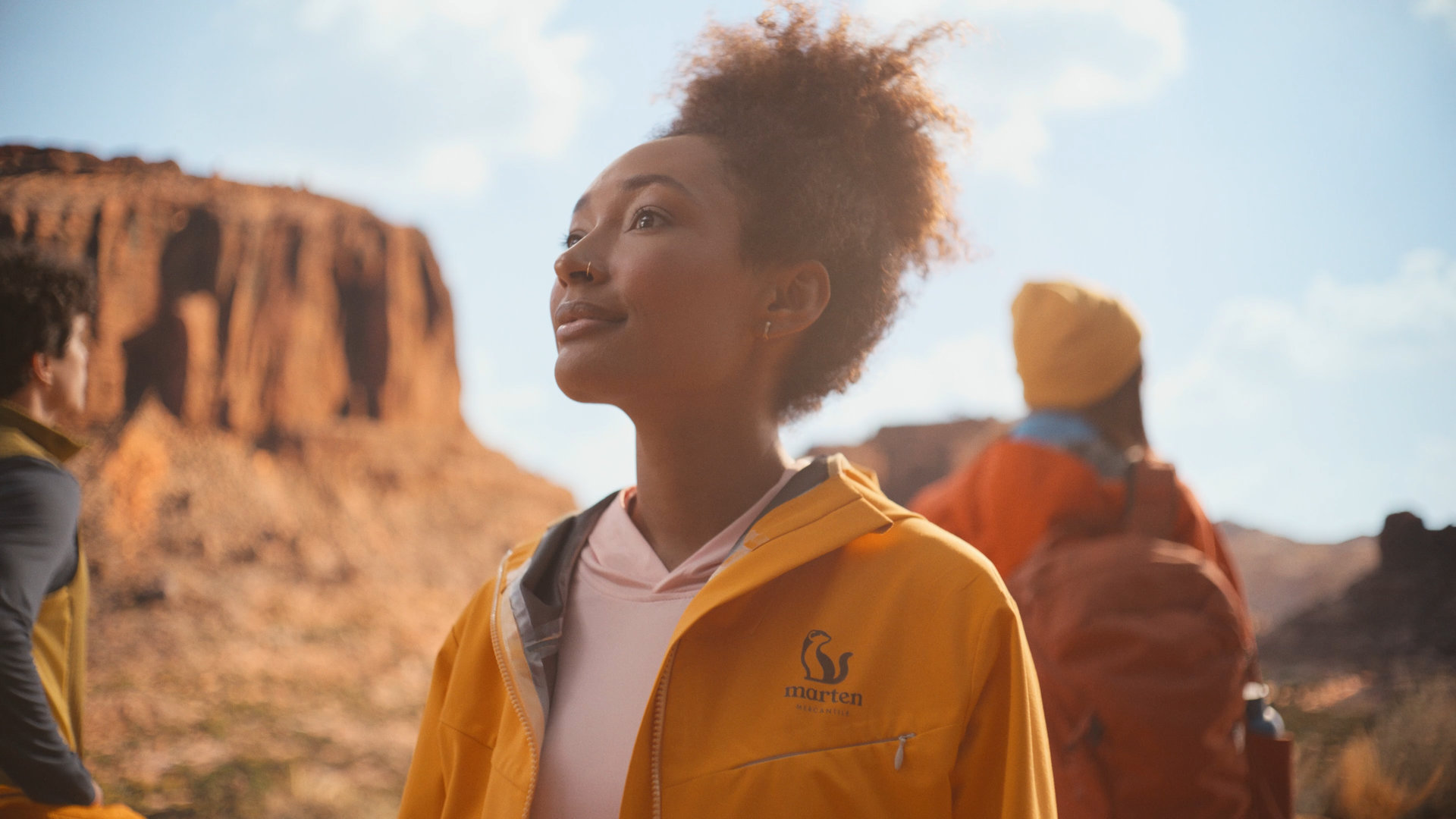 Three women standing outdoors in a desert landscape with red rock formations. The woman in the center has curly hair, a nose ring, and is wearing a yellow jacket with a logo on it, looking thoughtfully into the distance. The woman on the right has a yellow headscarf and an orange jacket, and the woman on the left has a dark jacket with lighter hair.